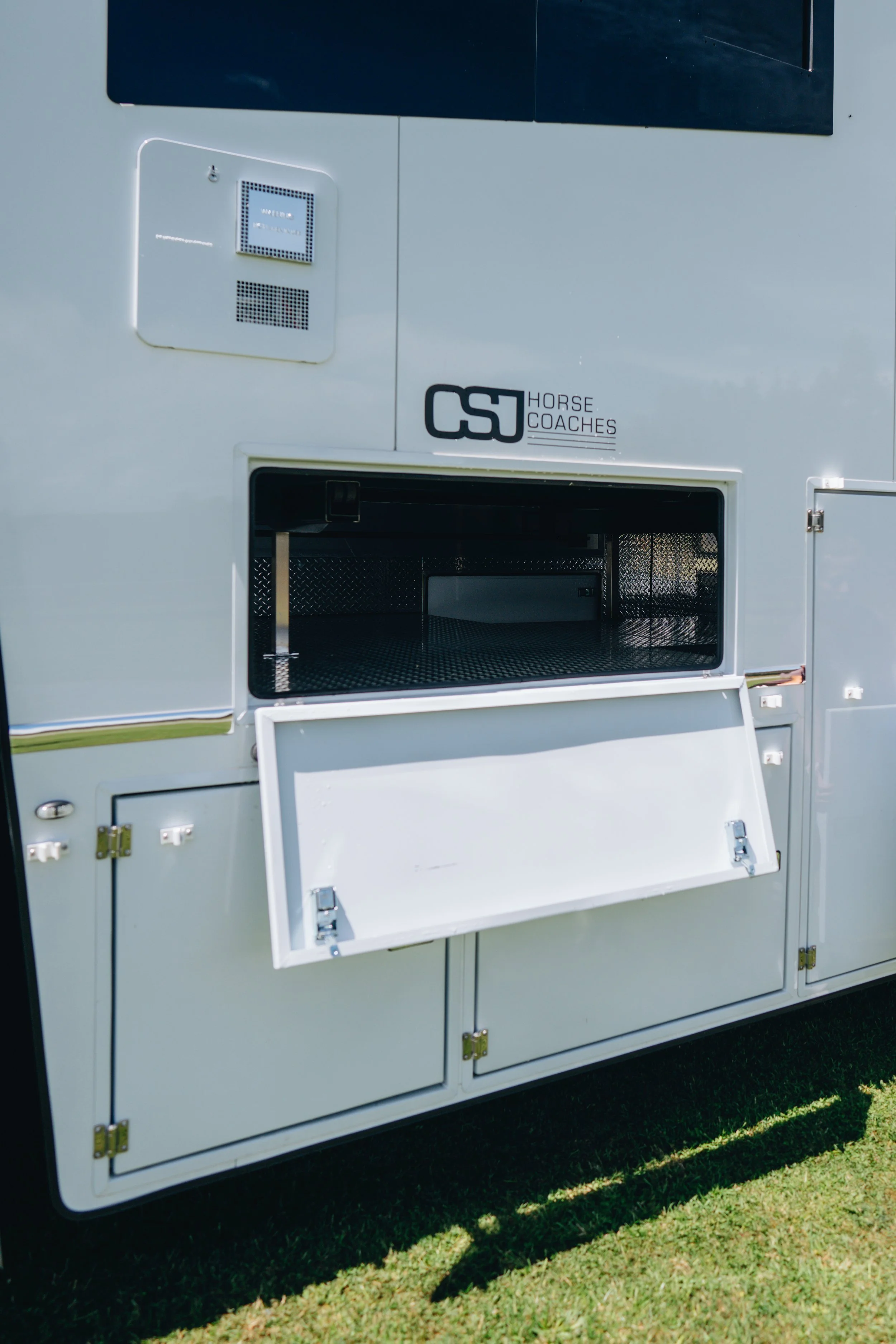A white horse trailer with a storage compartment door open, displaying a black metal interior. The trailer has a logo that reads 'CSJ Horse Coaches' on the side.