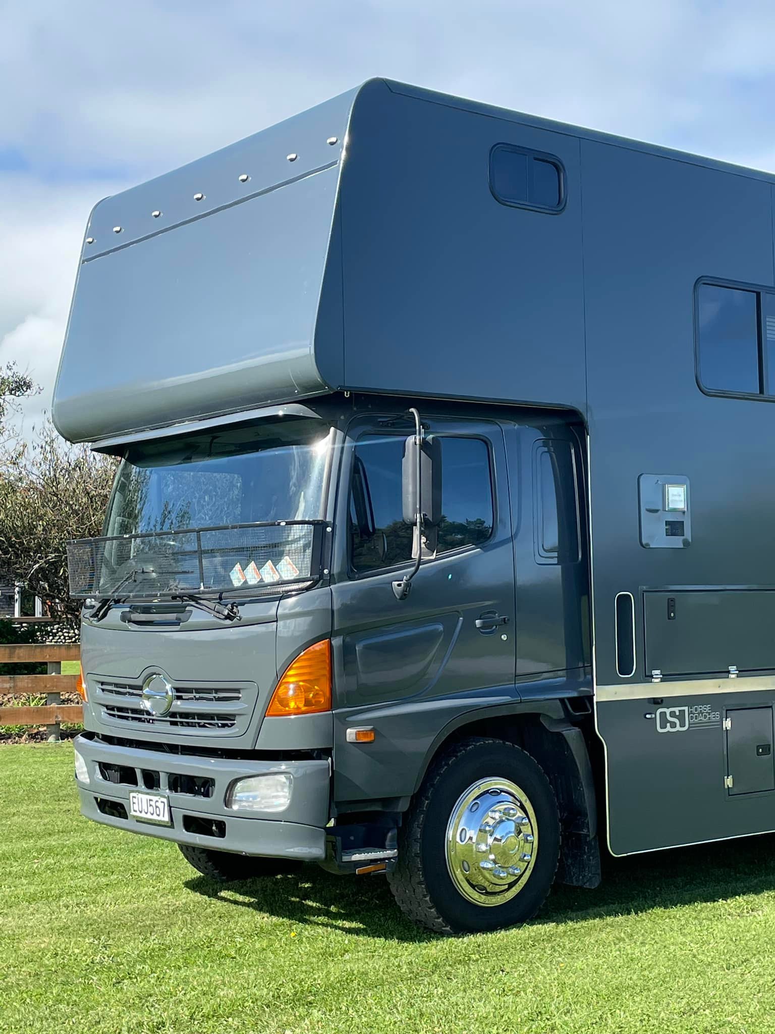 A large, dark gray mobile leisure vehicle with a cab-over design parked on green grass, with a wooden fence and some trees in the background.