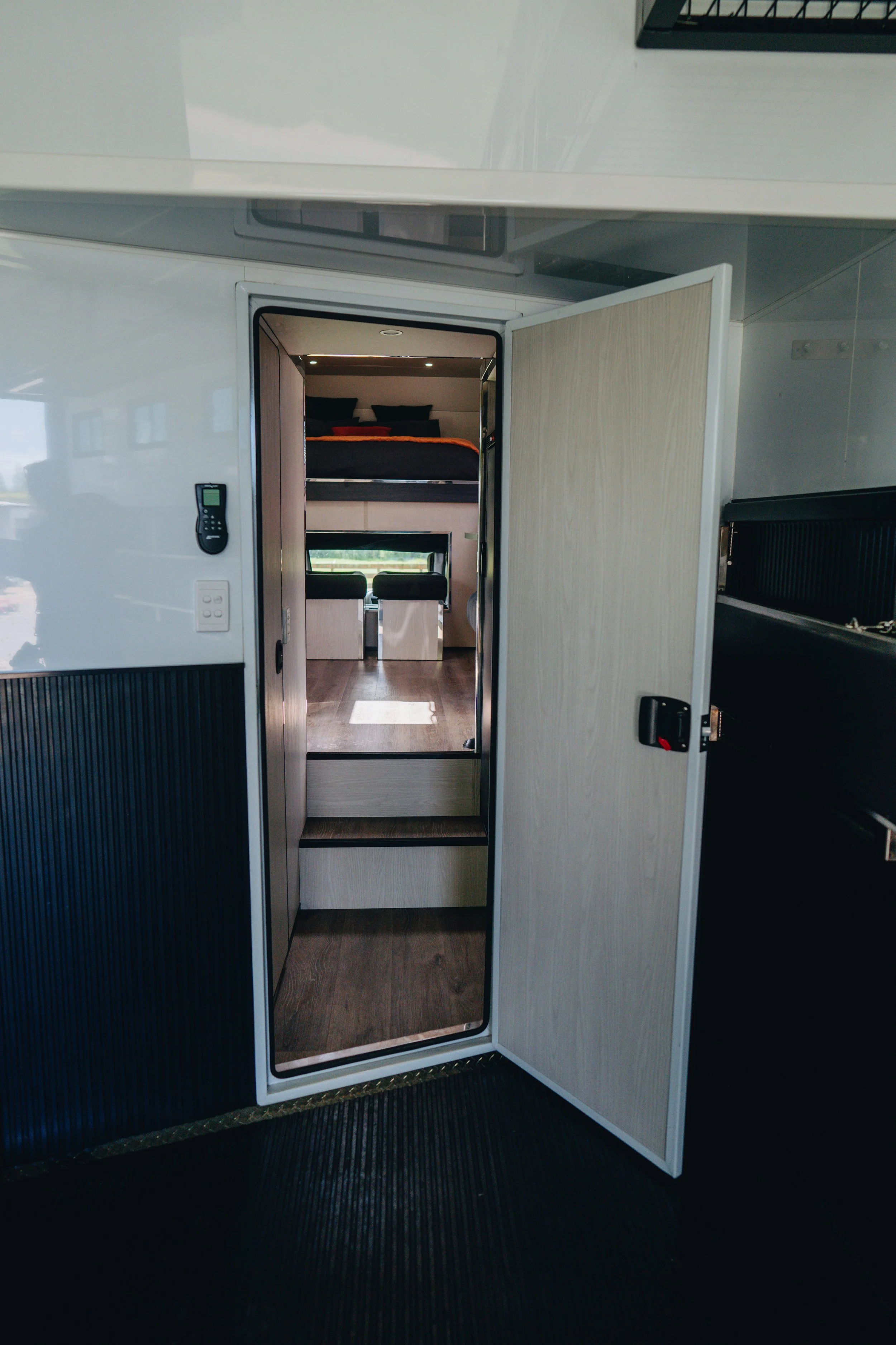 Open door leading to a modern RV interior with a bedroom, bed with pillows, living area with windows and seating.