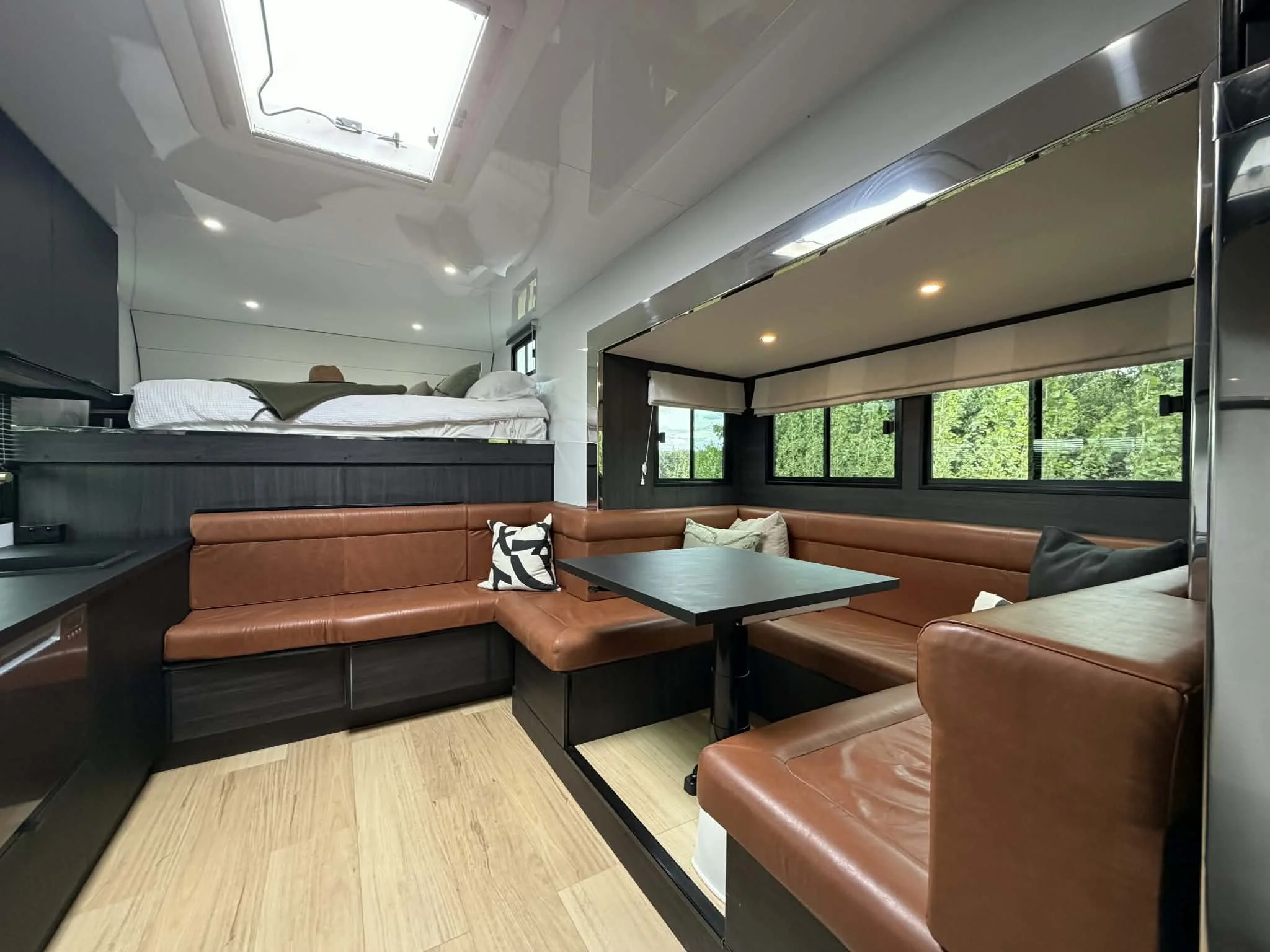 Interior of a modern compact living space with a cozy corner couch, a small black table, a bed elevated above, and windows showing greenery outside.