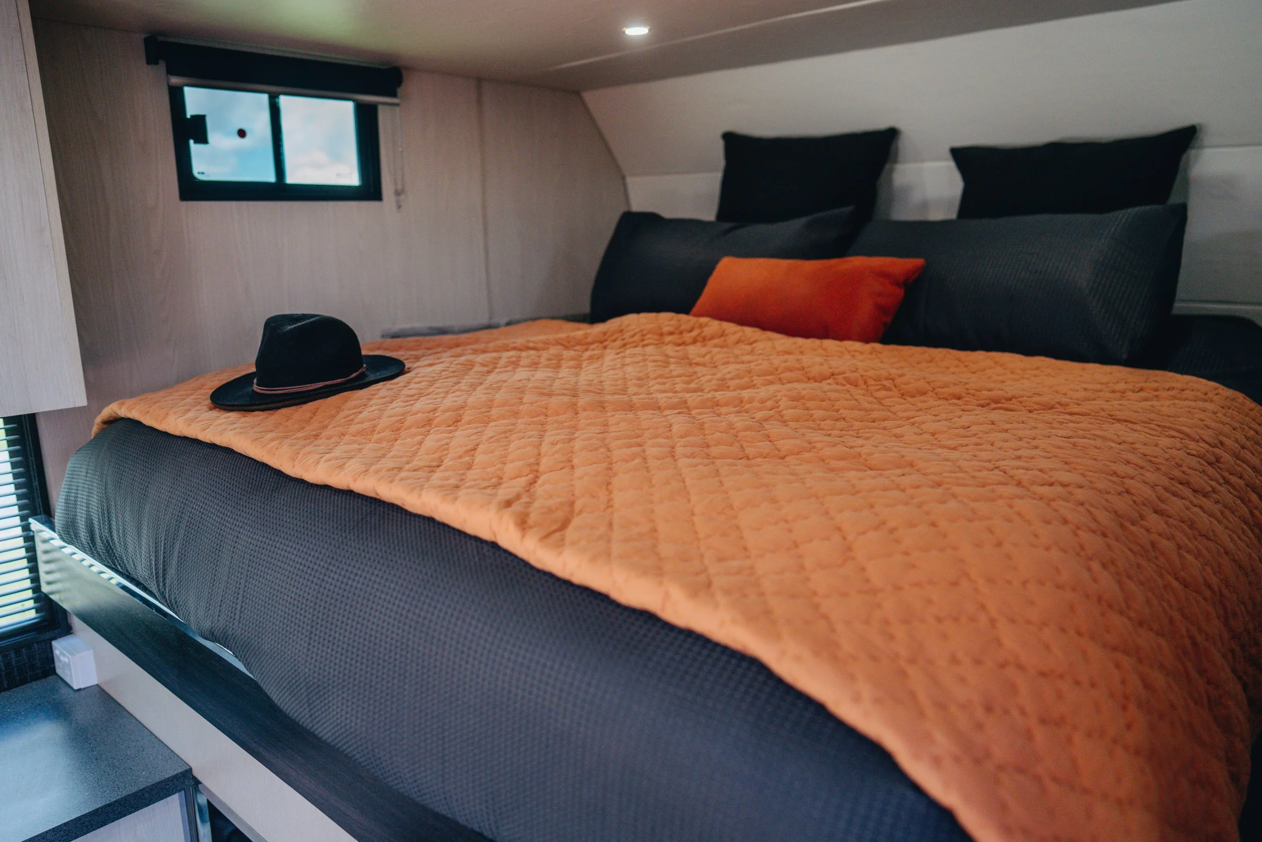 A neatly made bed with an orange quilt, black pillows, and a small orange pillow in a cozy room with a small window and natural lighting.