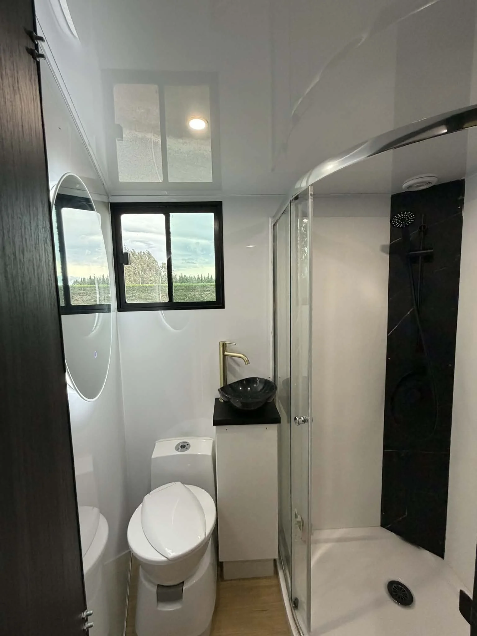 Small modern bathroom with a black sink, gold faucet, toilet, and glass shower enclosure. There is a window showing greenery outside.
