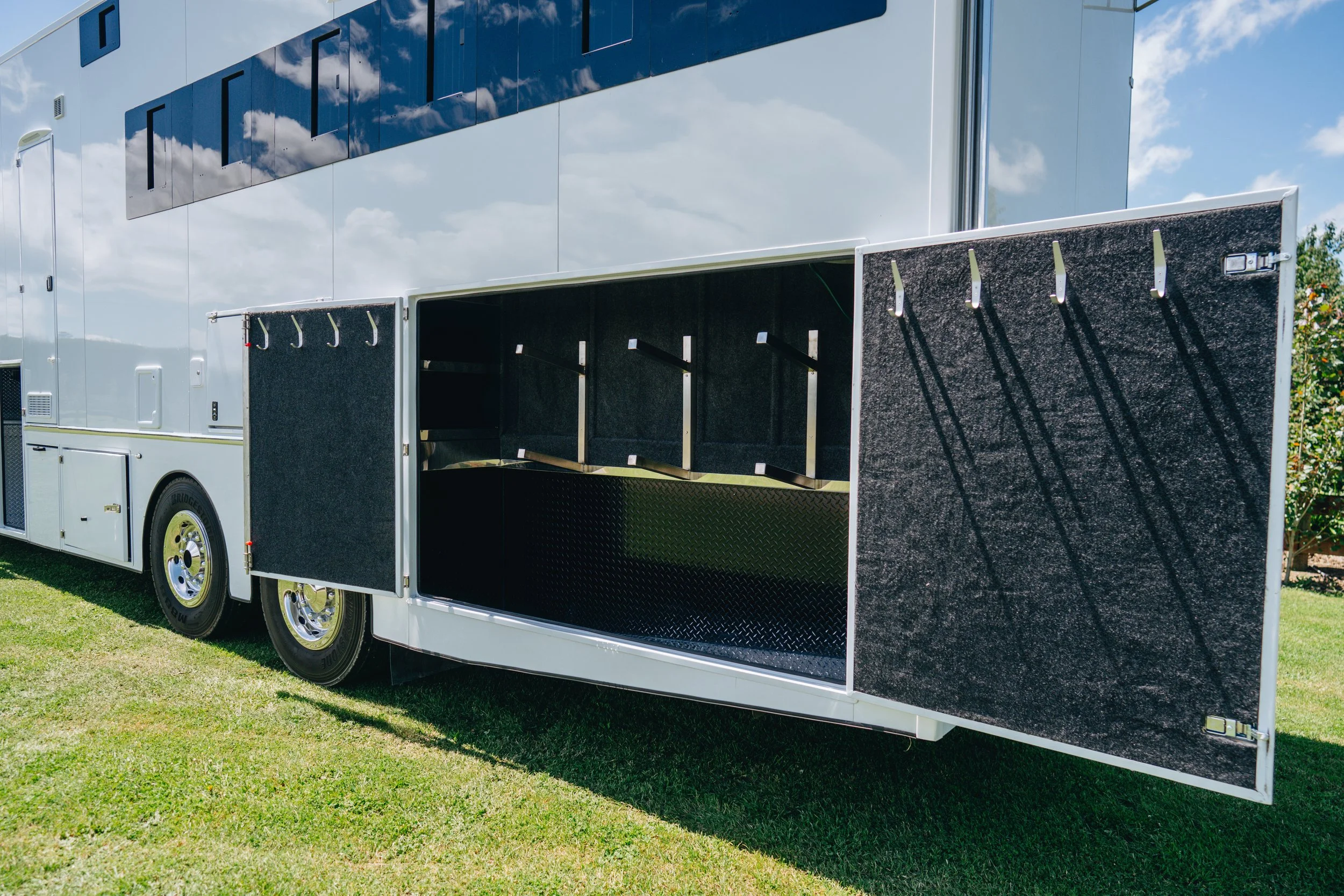 Open storage compartment of a large white utility truck with hooks and shelves, parked on a grassy area under a blue sky with scattered clouds.