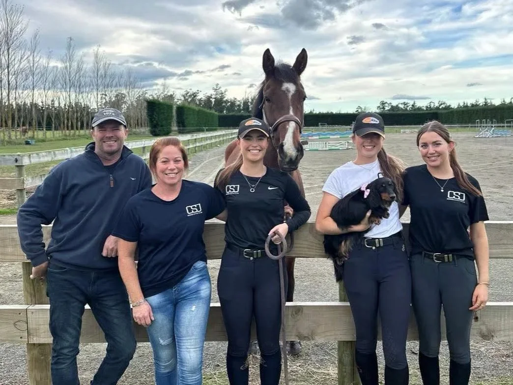 Group of five young people, three women and two men, posing with a horse and a small dog at an outdoor equestrian arena.