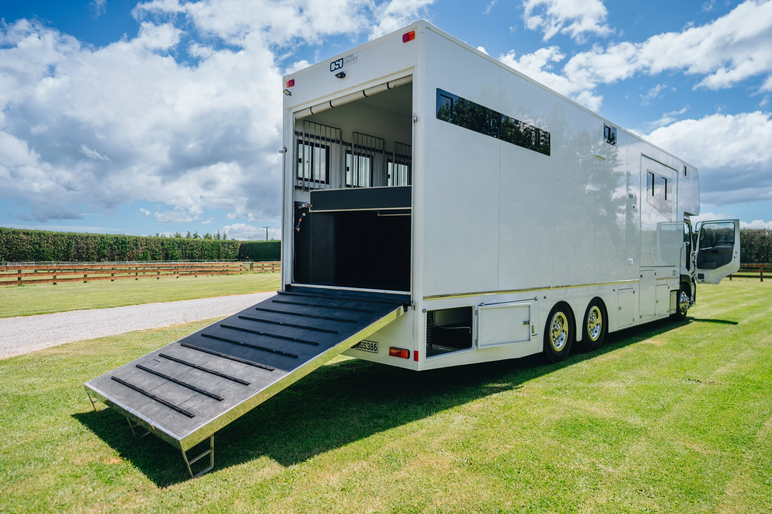 A white horse trailer with an extended ramp parked on a grassy field under a blue sky with scattered clouds.