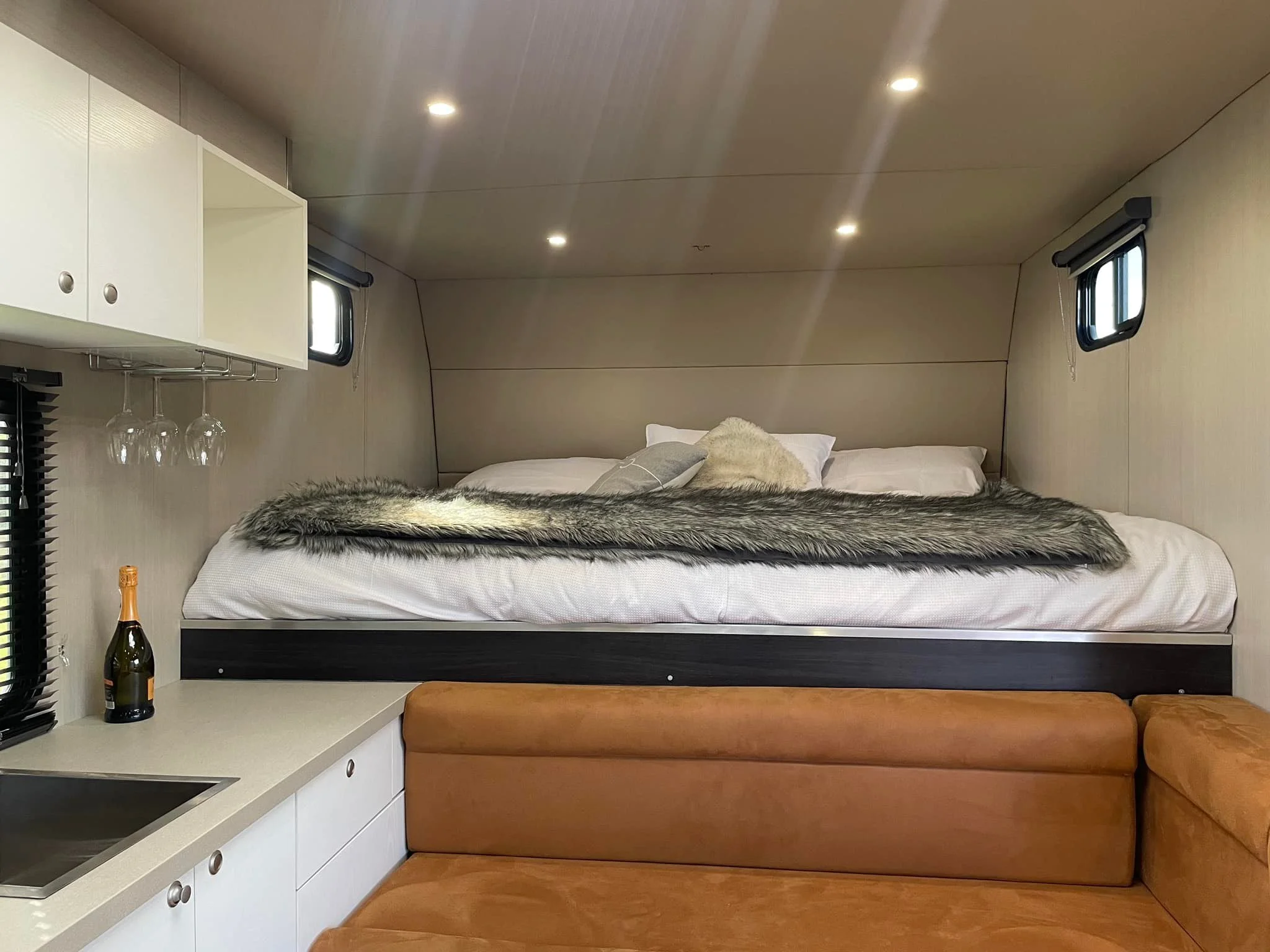 Interior of a cozy tiny home with a bed, fur throw, pillows, small windows, a counter with a bottle of champagne, wine glasses, and a sink.