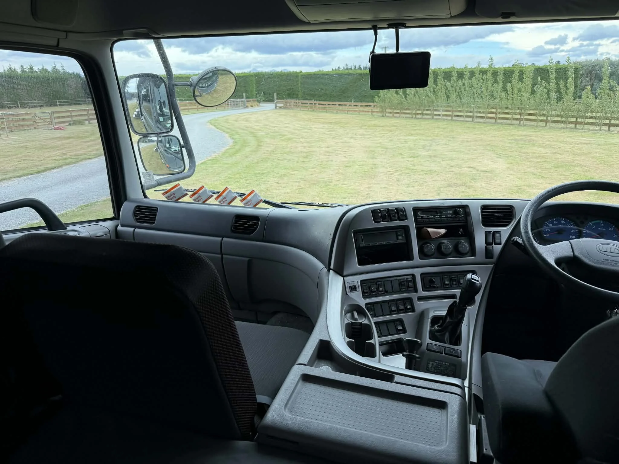 Interior of a bus cab with steering wheel, dashboard, gear shift, and side mirror. Outside view shows a curved gravel road, grassy field, and a hedge fence under cloudy skies.