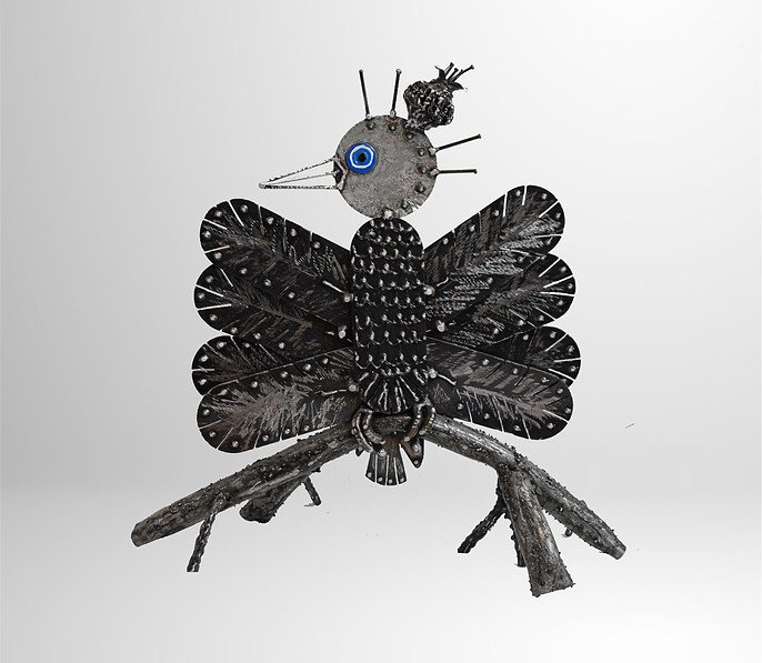 Sculpture of a bird made from metal and found objects, featuring a round body, large wings, and a distinct blue eye.