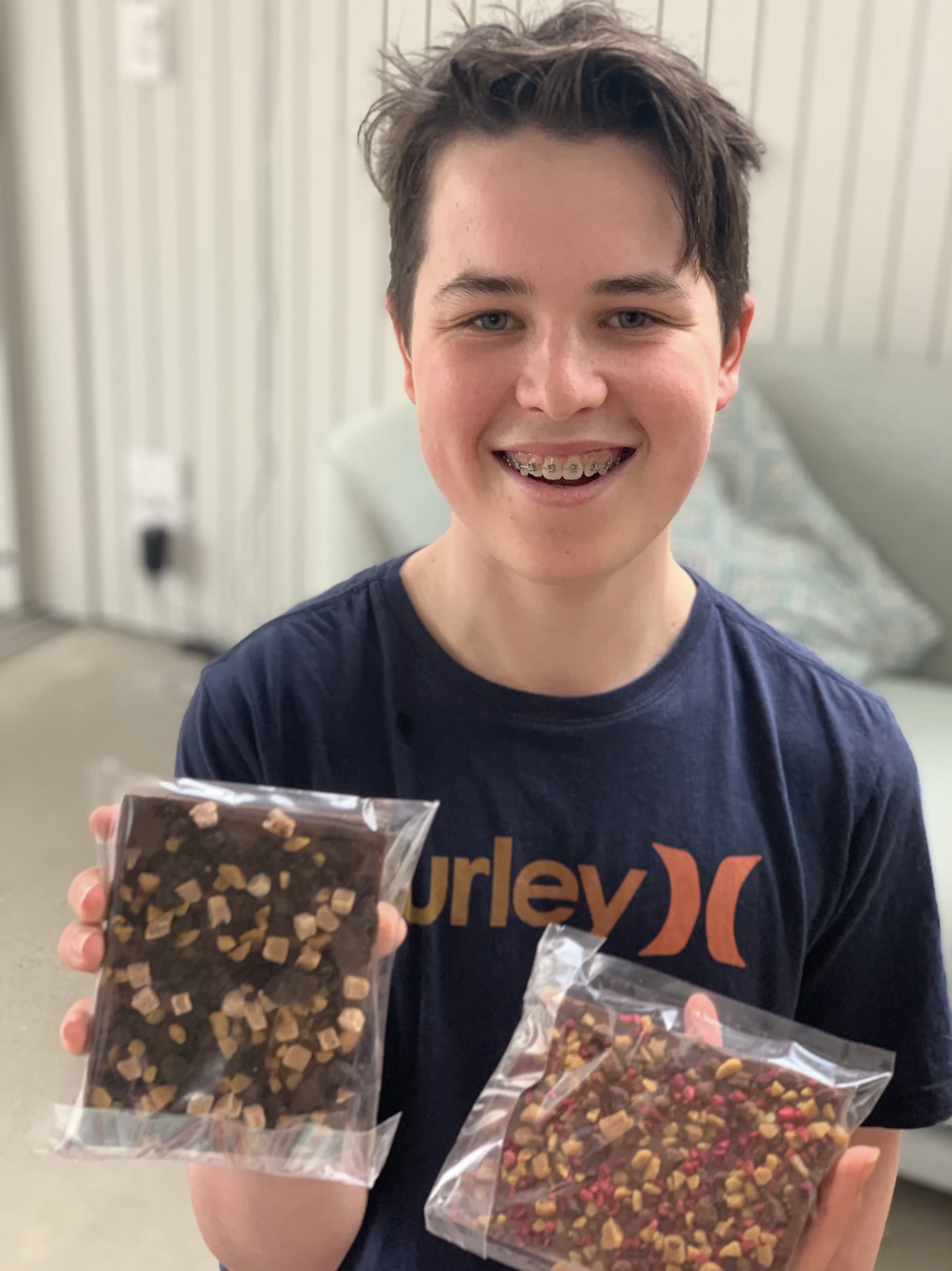 Young person with orthodontic braces smiling and holding two clear plastic bags of jar candies, standing in a room with white paneling and a bed in the background.