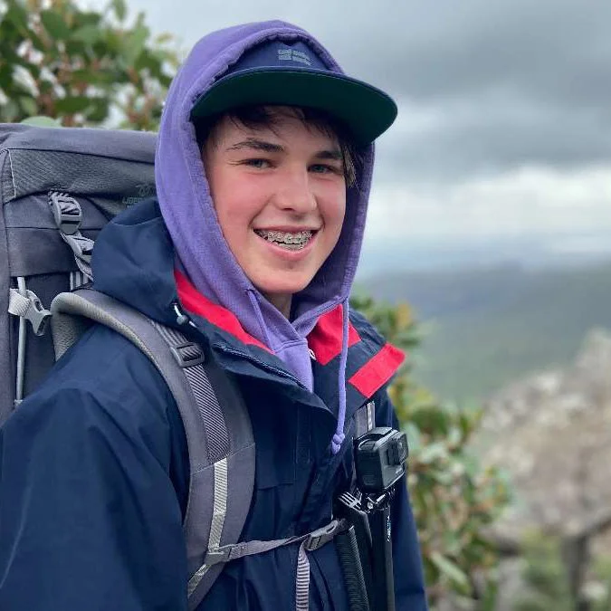Teenager outdoors in backpack and rain jacket, smiling, wearing a purple hoodie and black cap, with a mountainous landscape background.