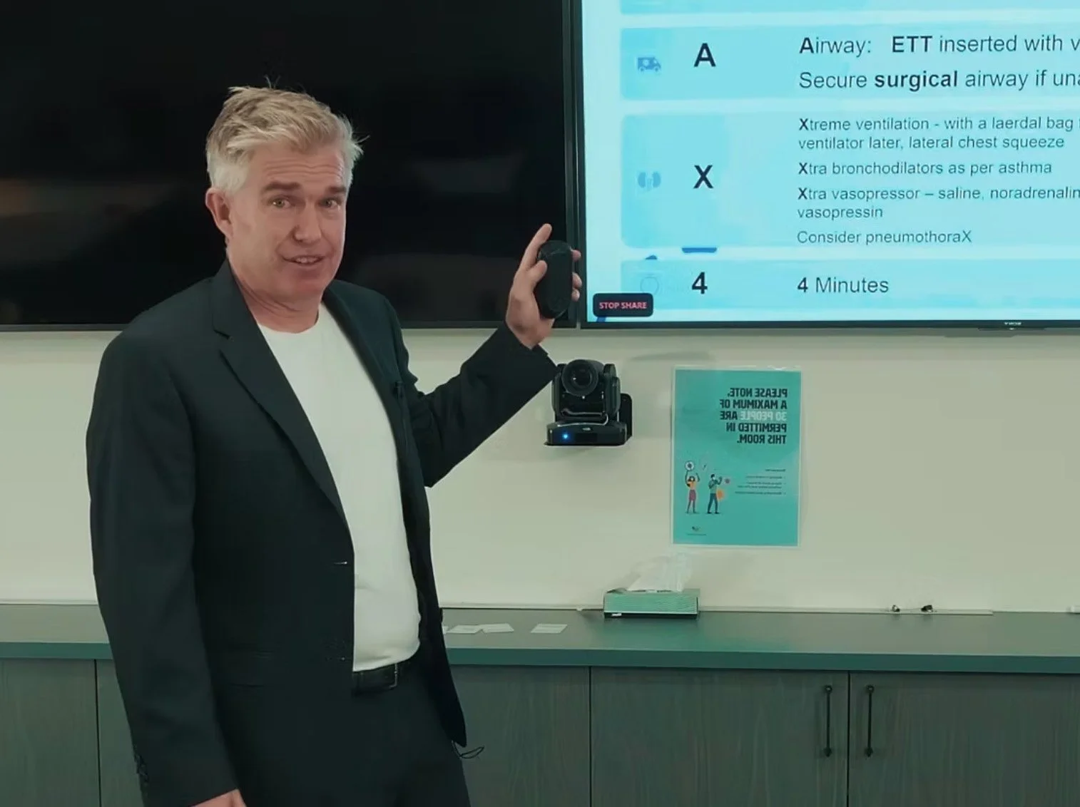 A man in a black blazer and white shirt standing in front of a large screen and pointing with a remote control. The screen displays medical instructions for airway management, including details about ET tube insertion and ventilation. A camera is mounted on the wall below the screen, and a poster with health information is also visible.