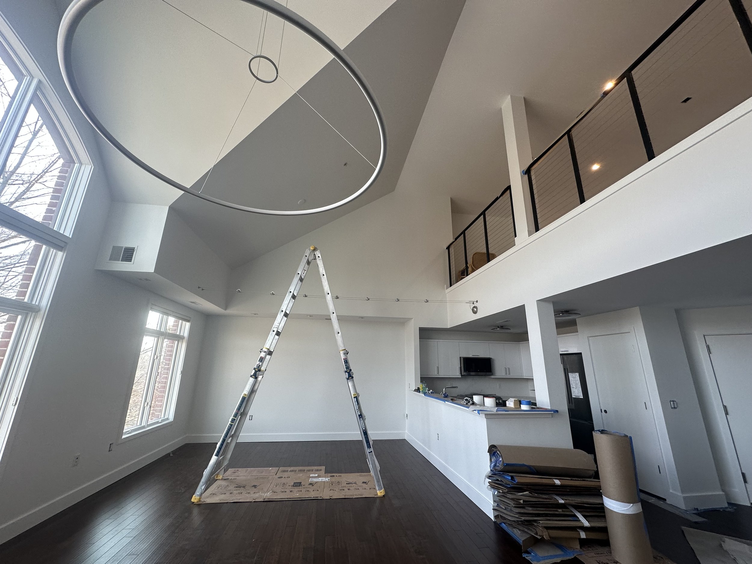 Empty living room with high ceiling, large windows, a ladder in the center, and kitchen in the background, indicating ongoing interior renovation.