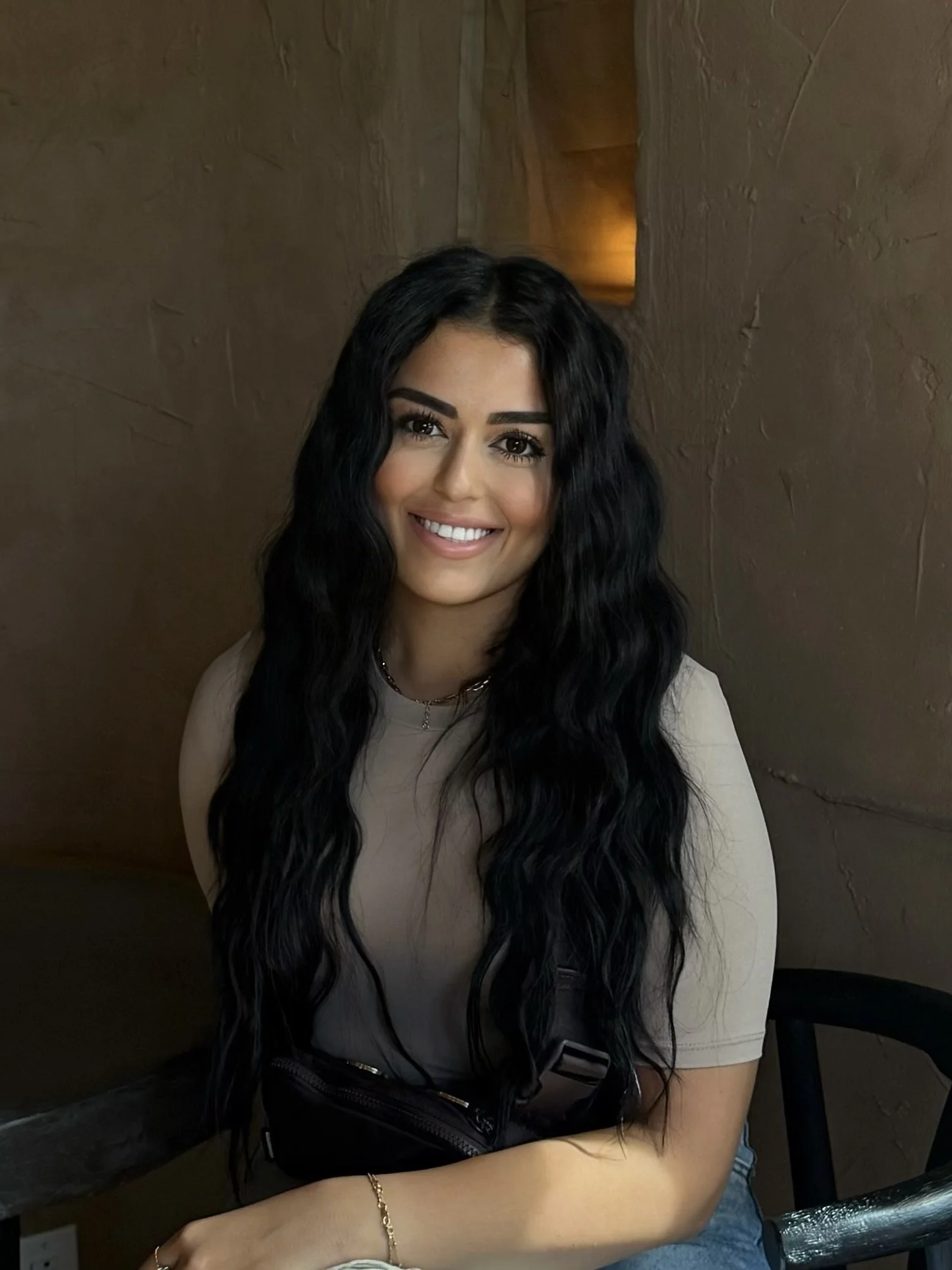 A smiling woman with long black wavy hair, wearing a beige top and sitting indoors against a textured brown wall.