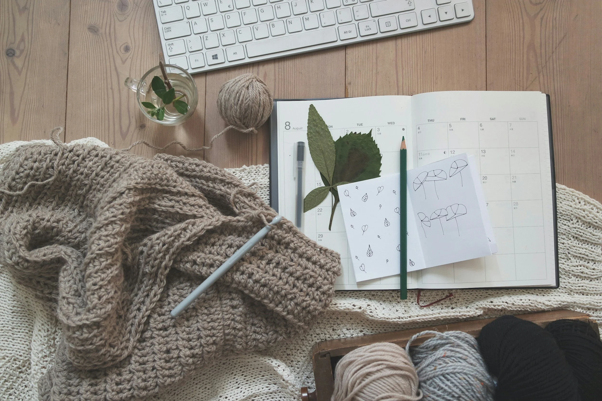 A flat lay of a cozy knitting workspace featuring a brown knitted blanket, a notebook with leafy and mushroom drawings, yarn balls in neutral and black colors, a glass of water with a plant, a ball of yarn, a white computer keyboard, and a piece of lined paper with leaf doodles.