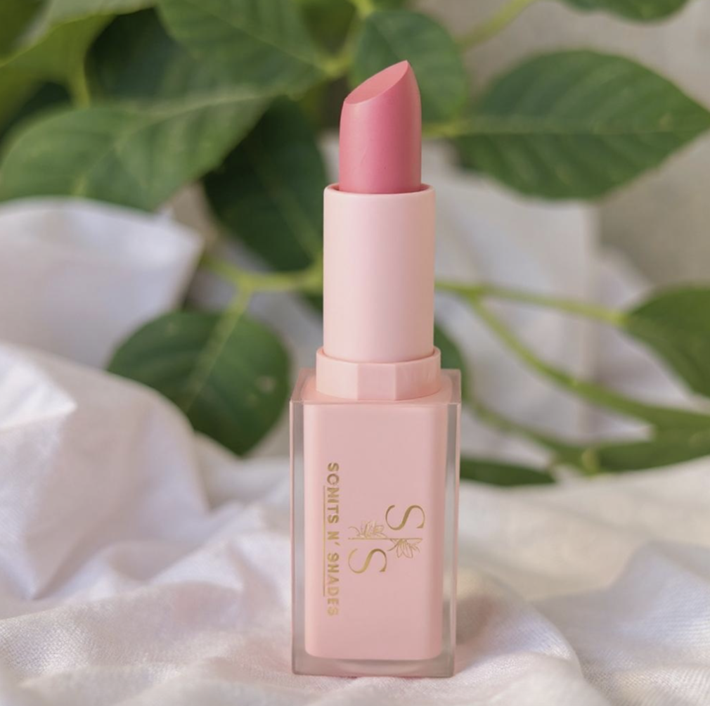 A pink lipstick in a light pink tube with gold lettering, placed on a soft white fabric with green leaves in the background.