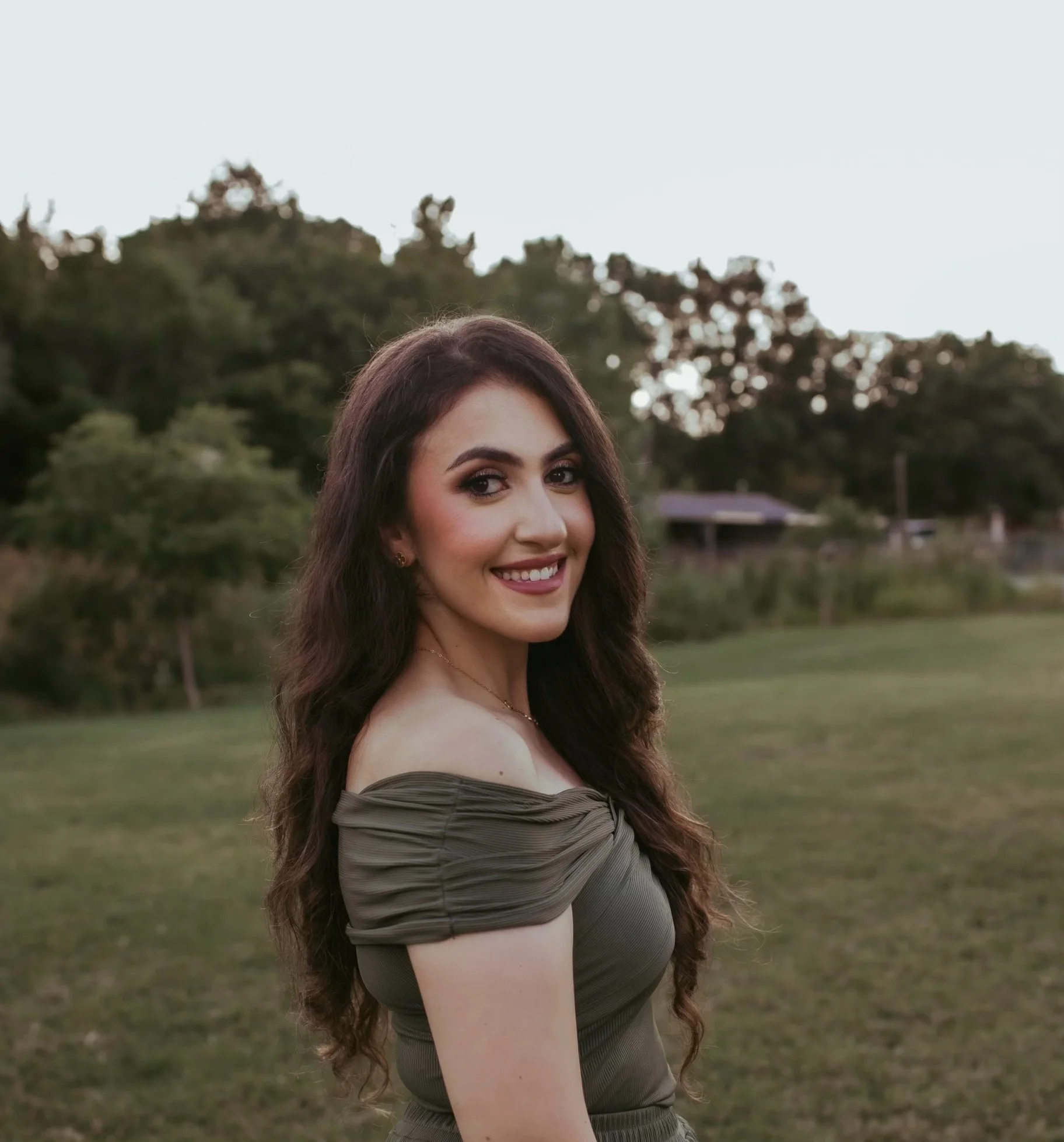 A young woman with long, wavy brown hair smiling outdoors during sunset, wearing an off-the-shoulder olive green dress, standing in a grassy field with trees in the background.