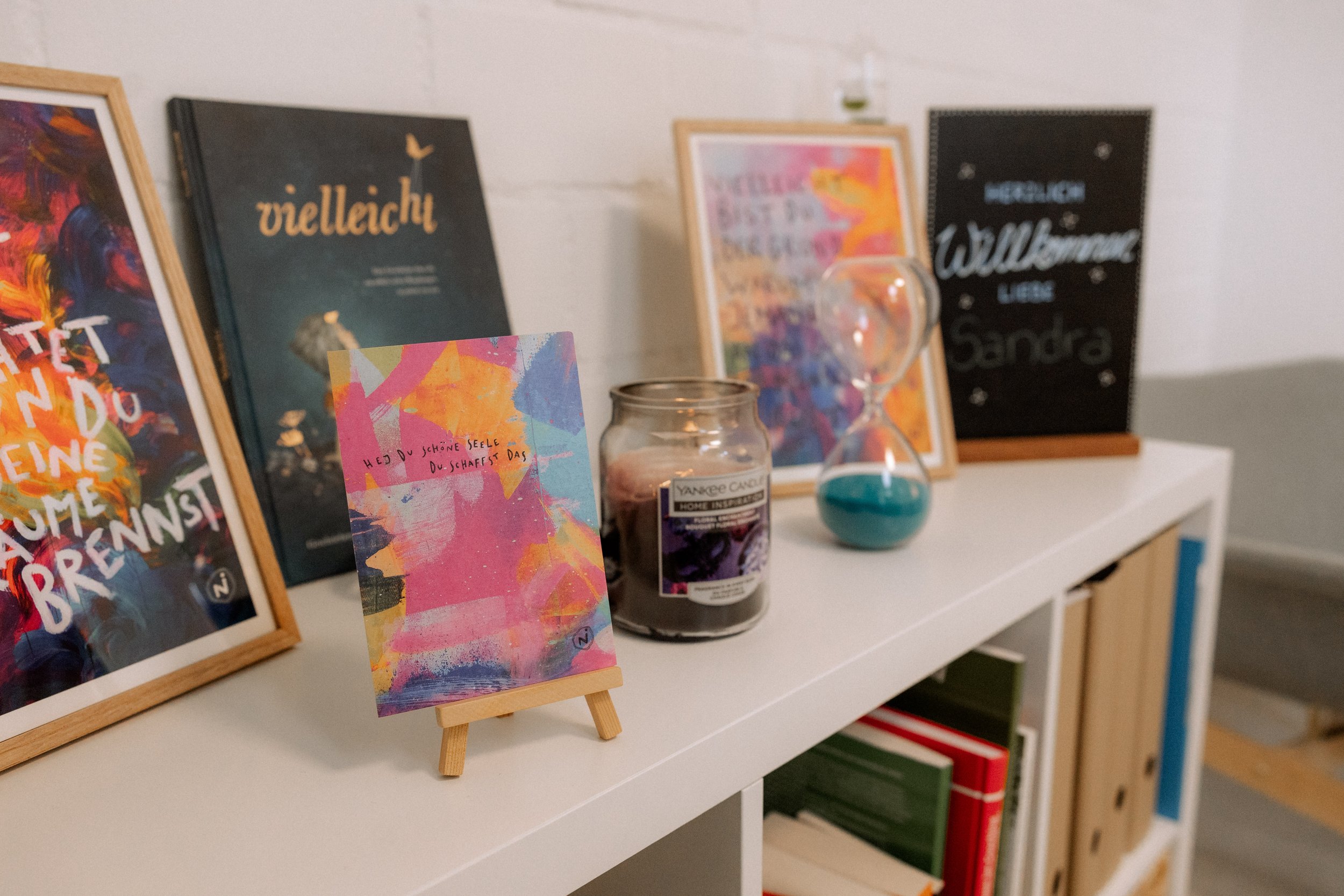 Bunte Bilder, Kerzen und Bücher auf einem weißen Regal