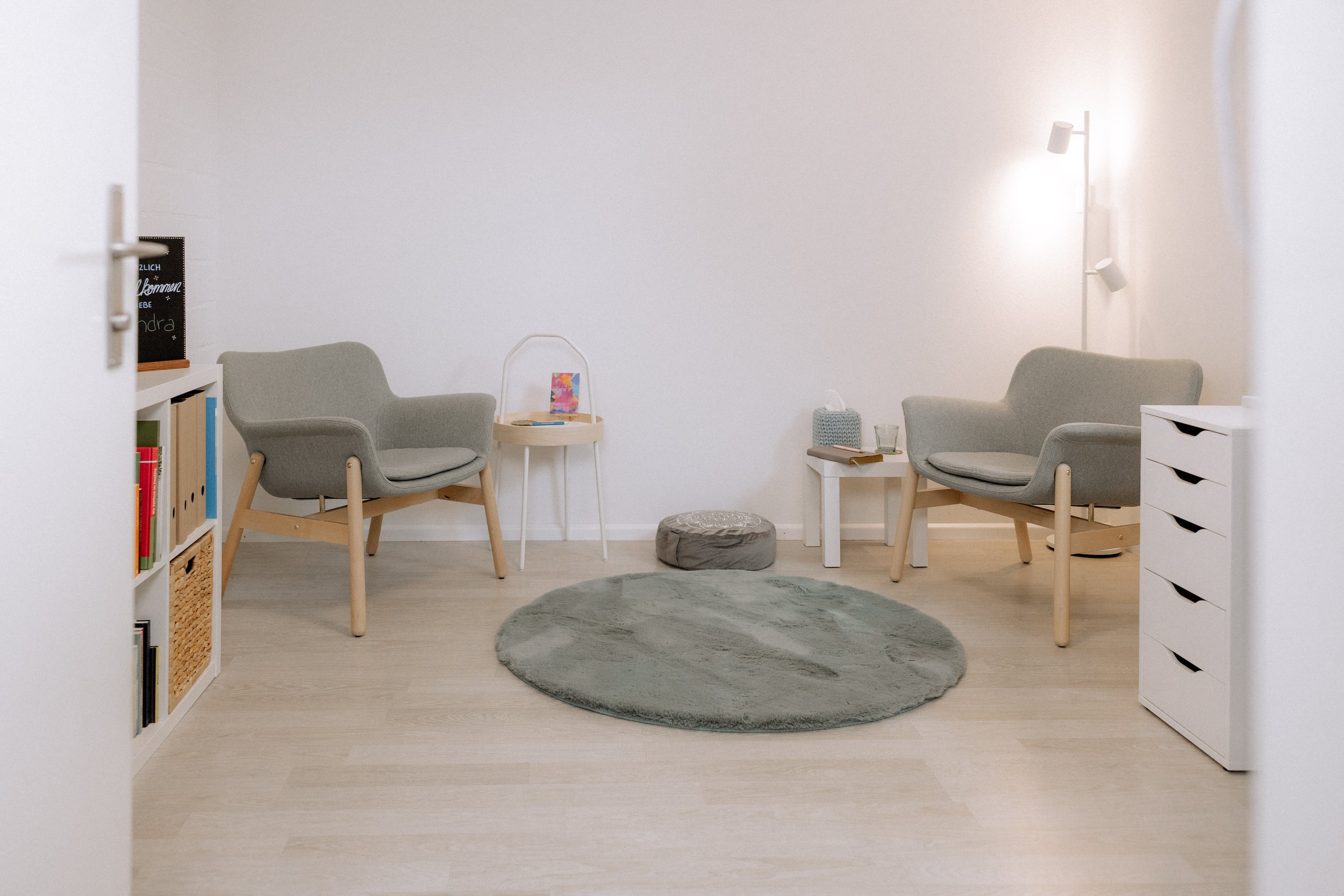 Gemütliches Wartezimmer mit zwei grauen Sesseln, kleinen Beistelltischen, einem runden Teppich, Stehlampe und weißen Möbeln in neutralen Tönen.