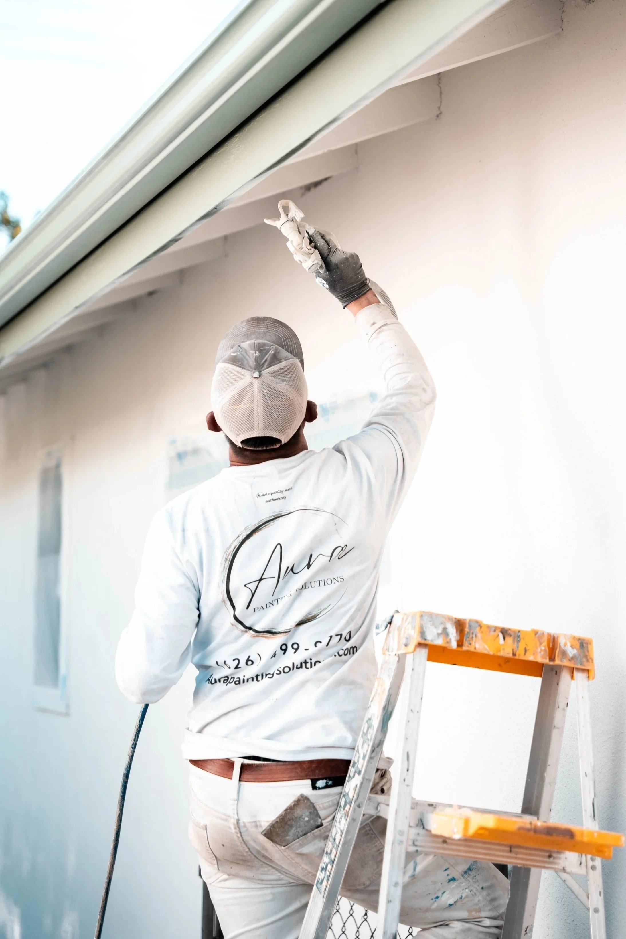 A painter is on a ladder, painting a white exterior wall of a house, wearing a cap, a long-sleeve shirt, and gloves.