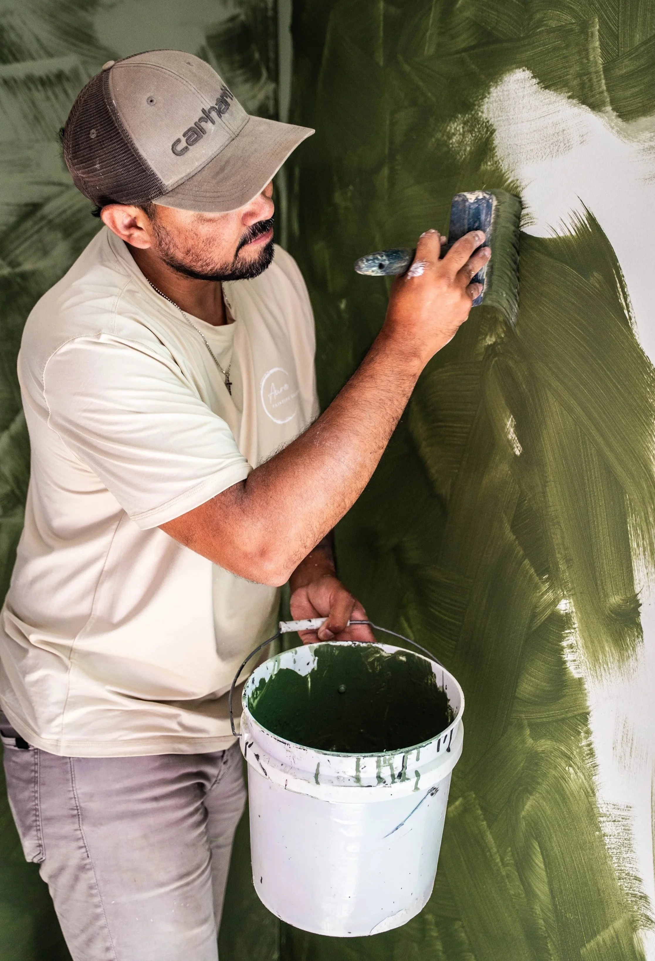 Man painting a green mural on a wall, holding a bucket of green paint in his left hand and applying paint with a roller with his right hand.