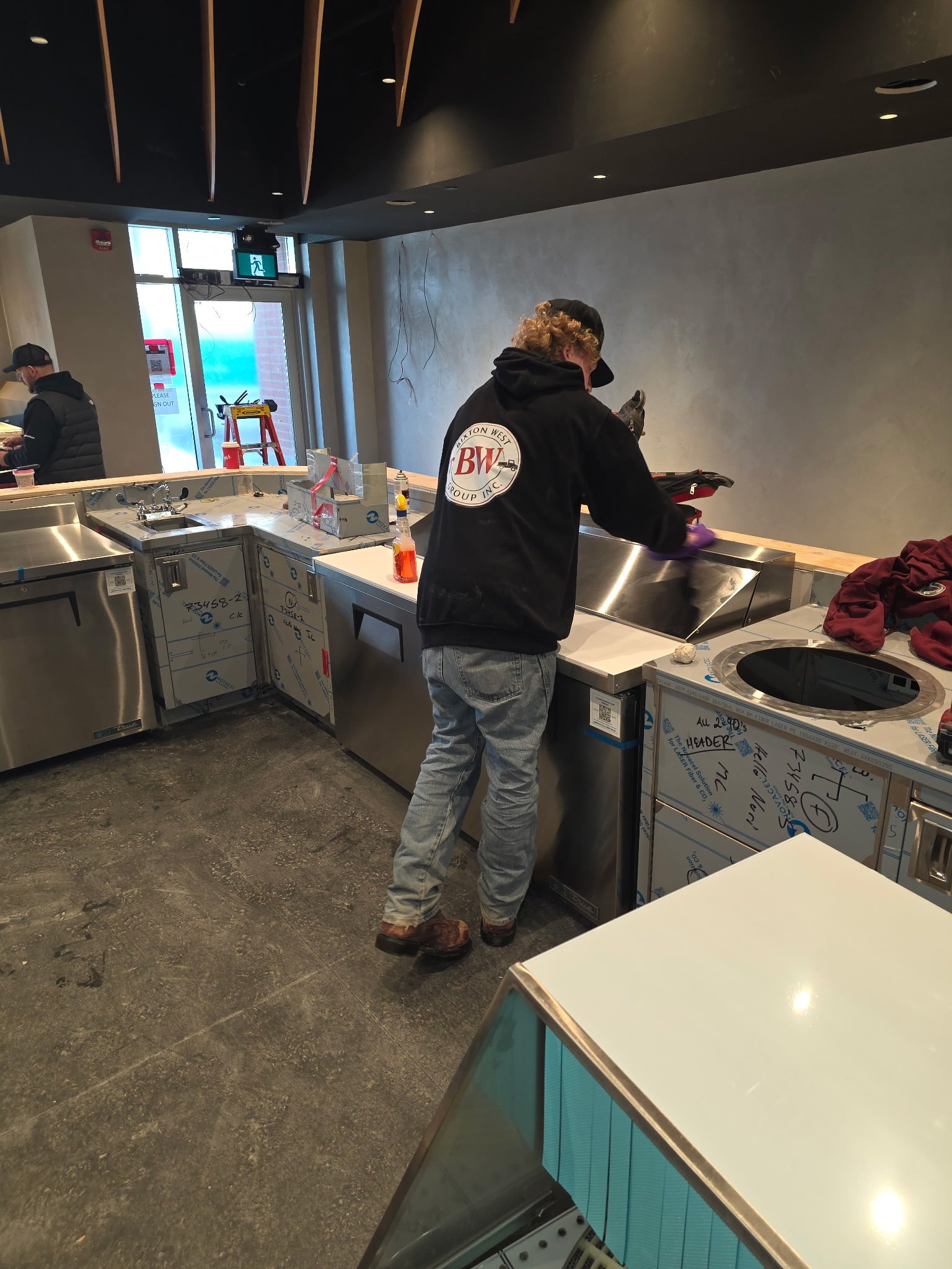 People working in a commercial kitchen or food service area. One person with curly hair is wearing a black hoodie with a logo on the back and gloves, cleaning or preparing the counter. Another person with a cap and vest is in the background. The space has a modern design with glossy counters, some boxes, and cleaning supplies. There is an emergency exit sign above the door.