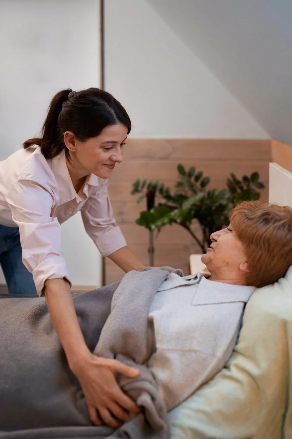 A caregiver leaning over a patient in bed, holding their hand and smiling at them.