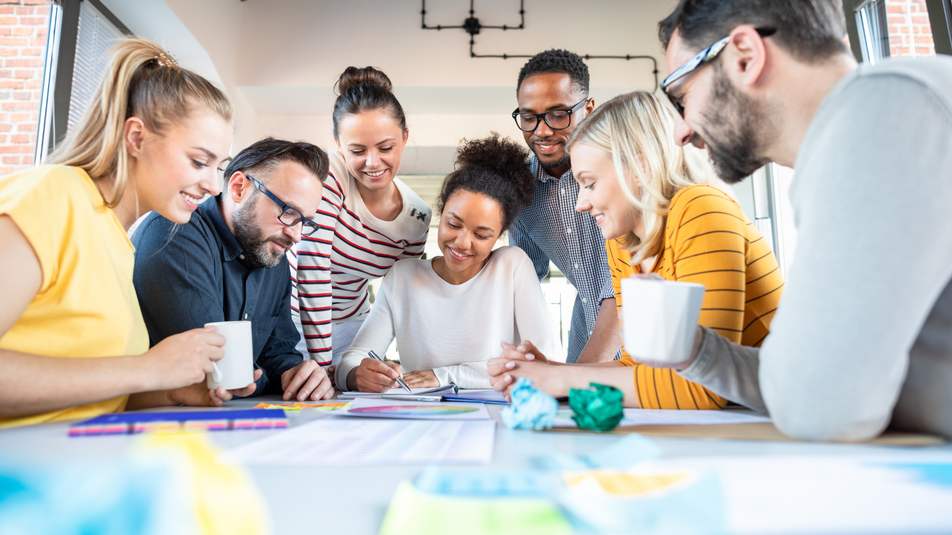 Group of eight diverse people gathered around a table, smiling, and working together, with paper, notebooks, and coffee mugs in a bright office.