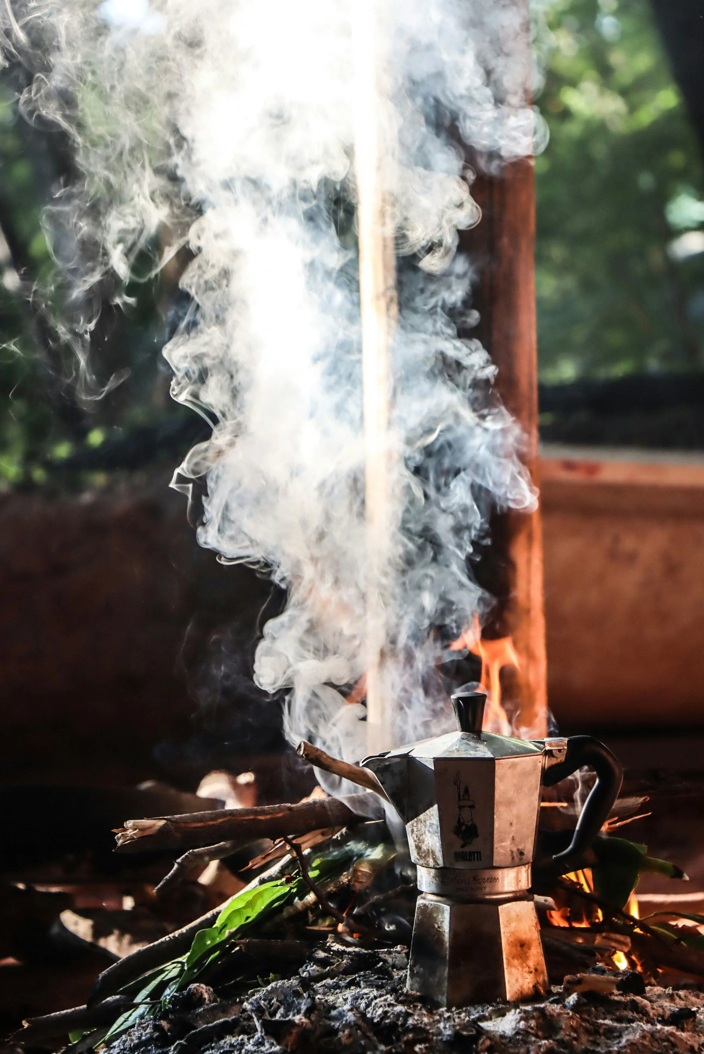 A stovetop tea kettle on an open fire surrounded by sticks and leaves, with smoke rising from it.