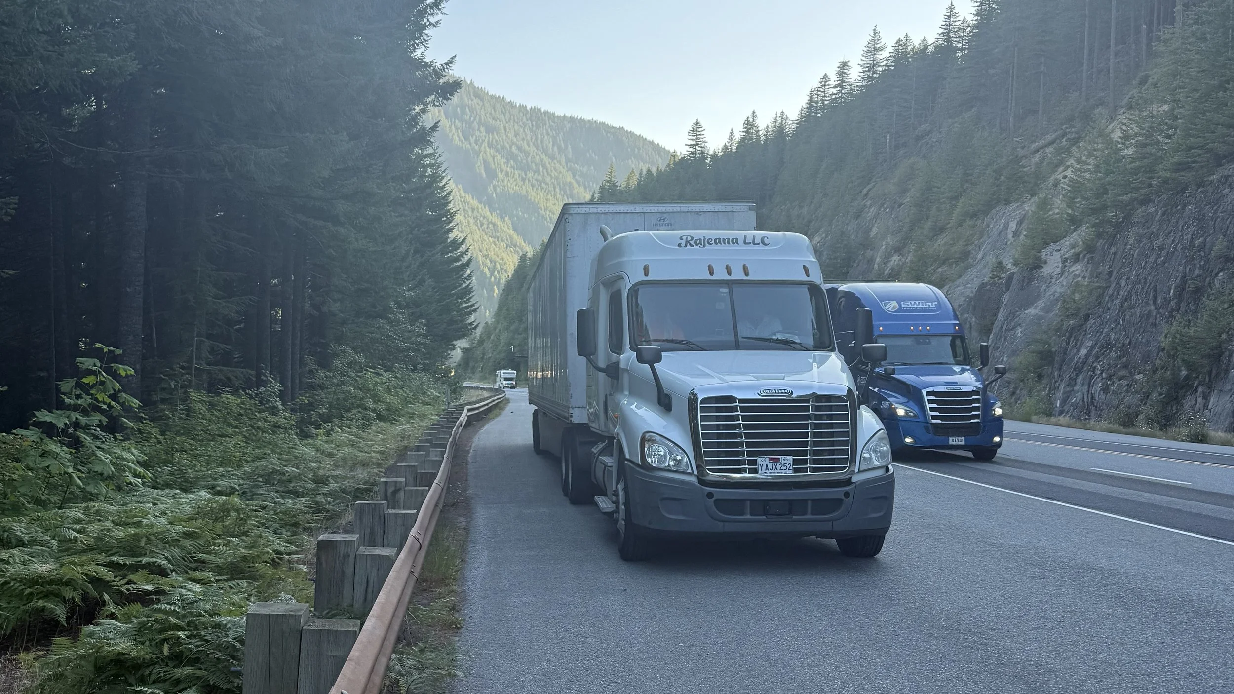 Two semi-trucks parked on the shoulder of a mountain road, surrounded by dense evergreen forest and rocky hillsides.