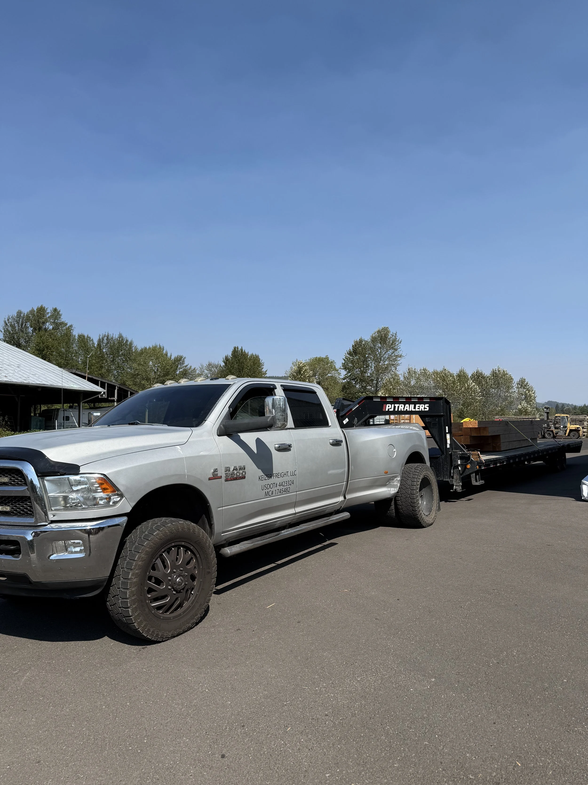Silver RAM truck with a flatbed trailer carrying wooden planks next to a building and trees under a clear blue sky.