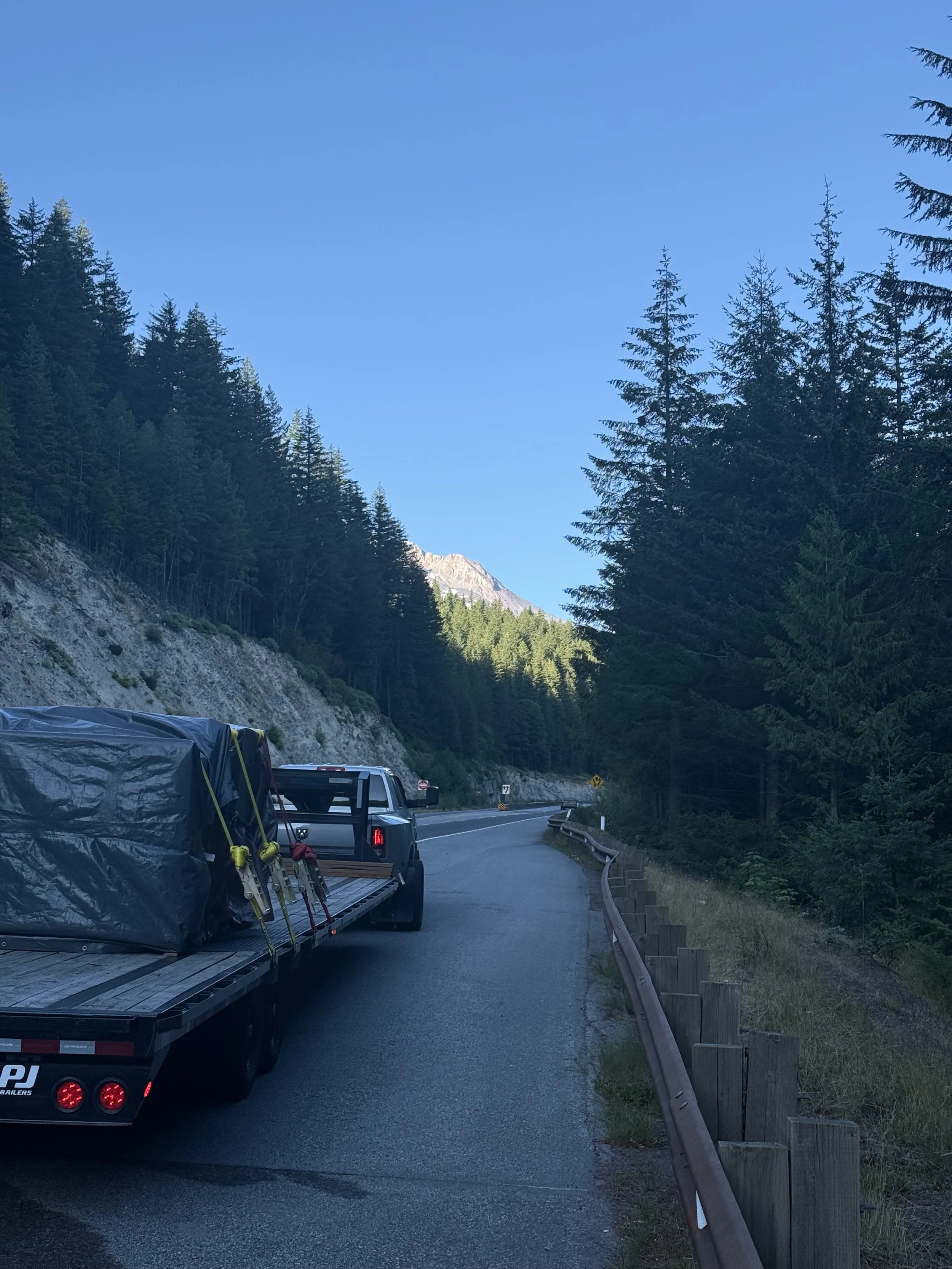 A black pickup truck towing a flatbed trailer carrying camping gear, driving on a winding mountain road lined with trees and guardrails under a clear blue sky.