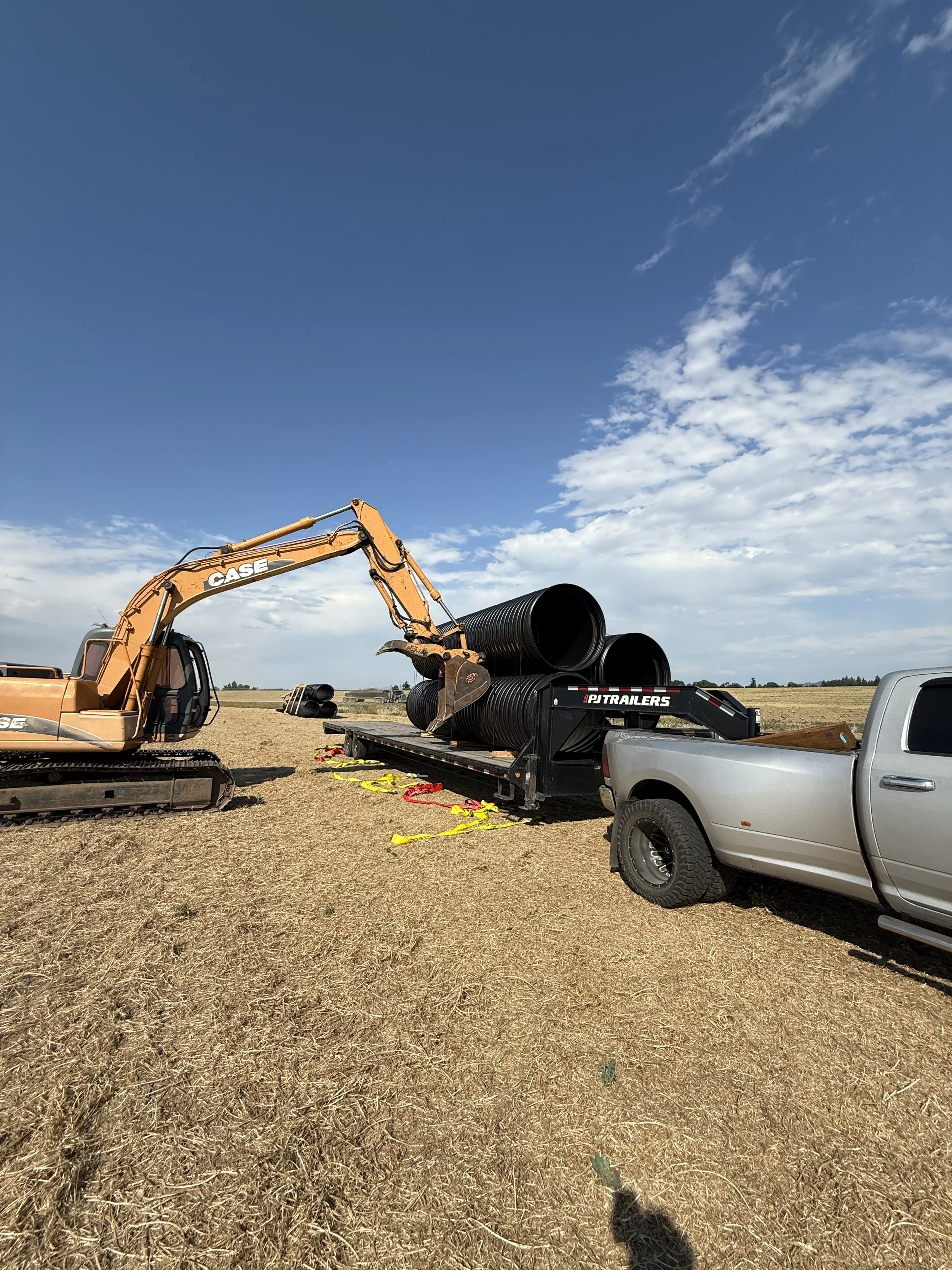 A construction site in an open field with a large orange excavator loading big black pipes onto a flatbed trailer attached to a silver pickup truck, under a partly cloudy sky.