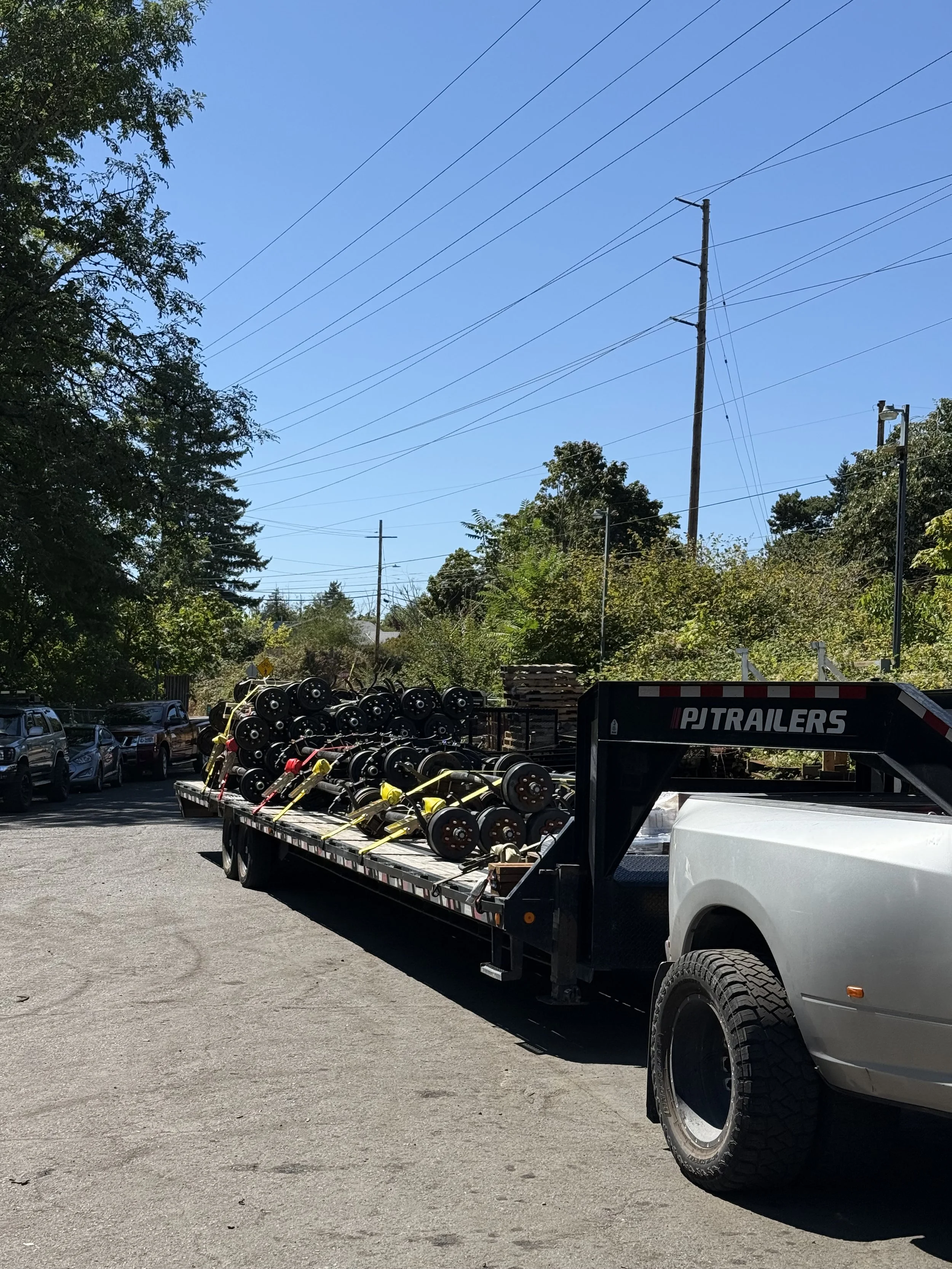 Flatbed truck carrying multiple black wheels or rollers with yellow straps, parked in a lot with other vehicles and trees in the background under a clear blue sky.