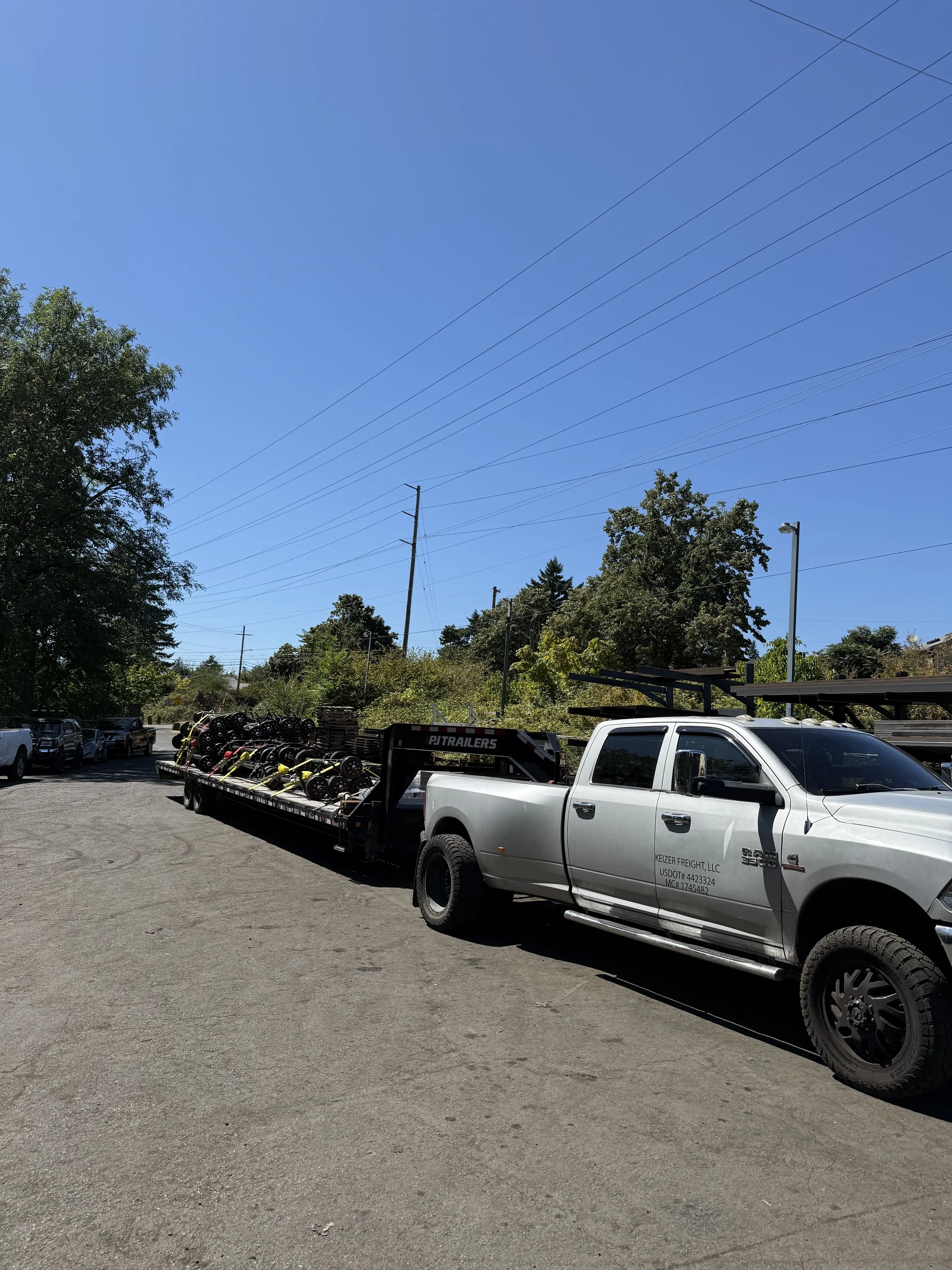 A silver pickup truck with a flatbed trailer carrying various equipment, parked on a paved lot under a clear blue sky, with trees and utility poles in the background.