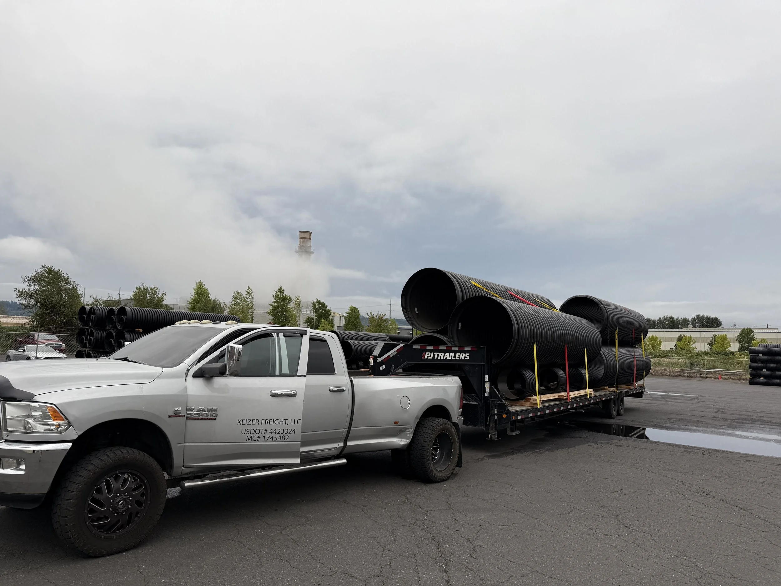 Silver pickup truck carrying large black drainage pipes parked in an outdoor lot with industrial buildings and smoke or steam rising in the distance.