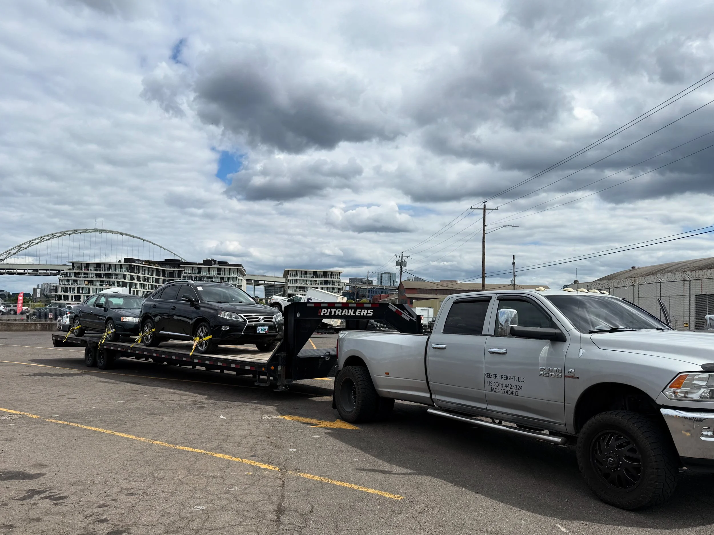 A white pickup truck towing a flatbed trailer carrying two black SUVs, parked in an outdoor lot with cloudy sky and cityscape in the background.