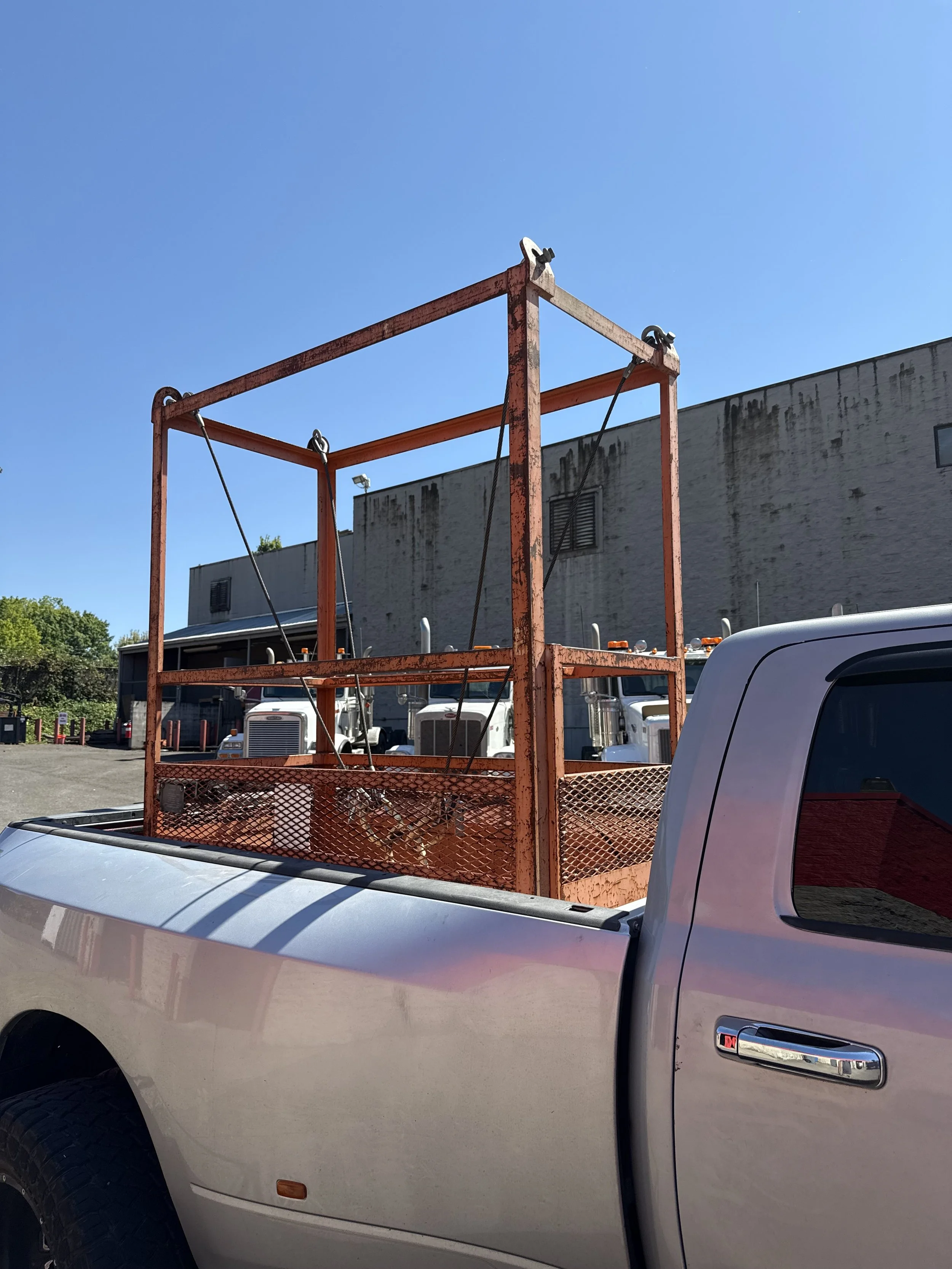 Silver pickup truck with a rusted orange metal cage in the truck bed, parked in a lot with trucks in the background, against a clear blue sky.