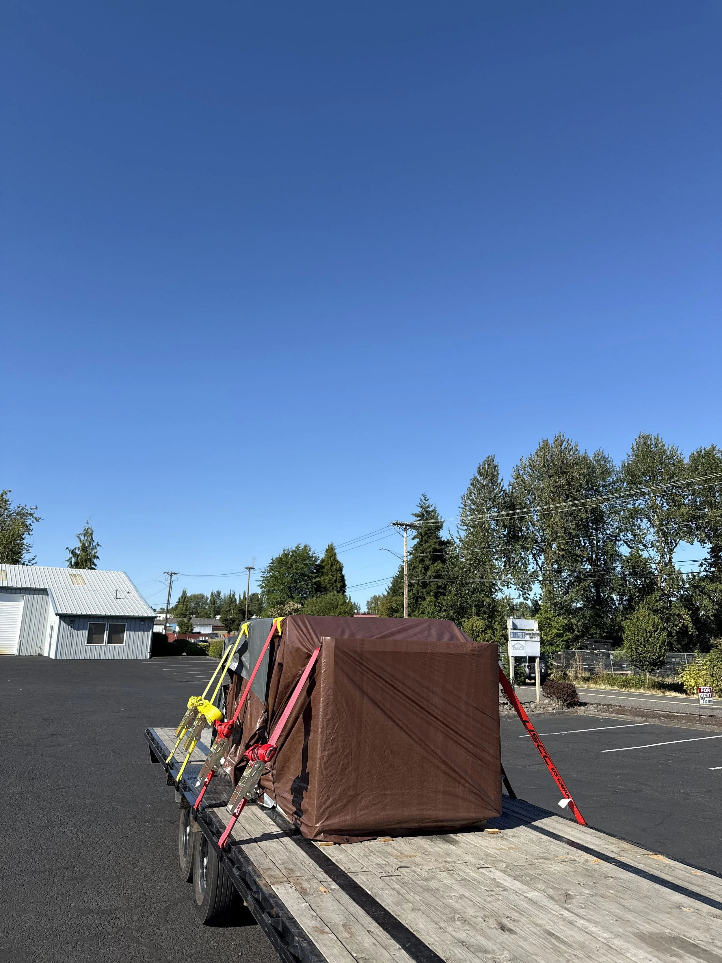 A flatbed trailer with a brown covered cargo and red and yellow straps holding it down, parked on a paved lot under a clear blue sky with some trees and a small building in the background.