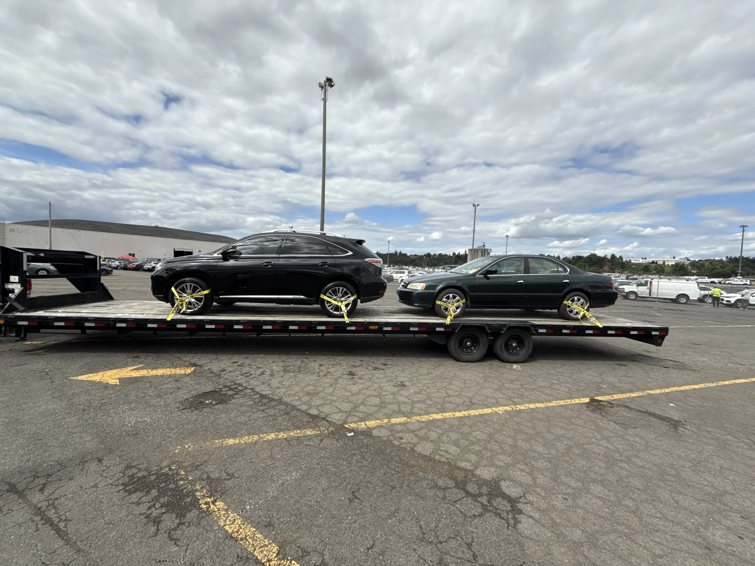 Two black cars secured on a flatbed trailer in a parking lot, under a cloudy sky.