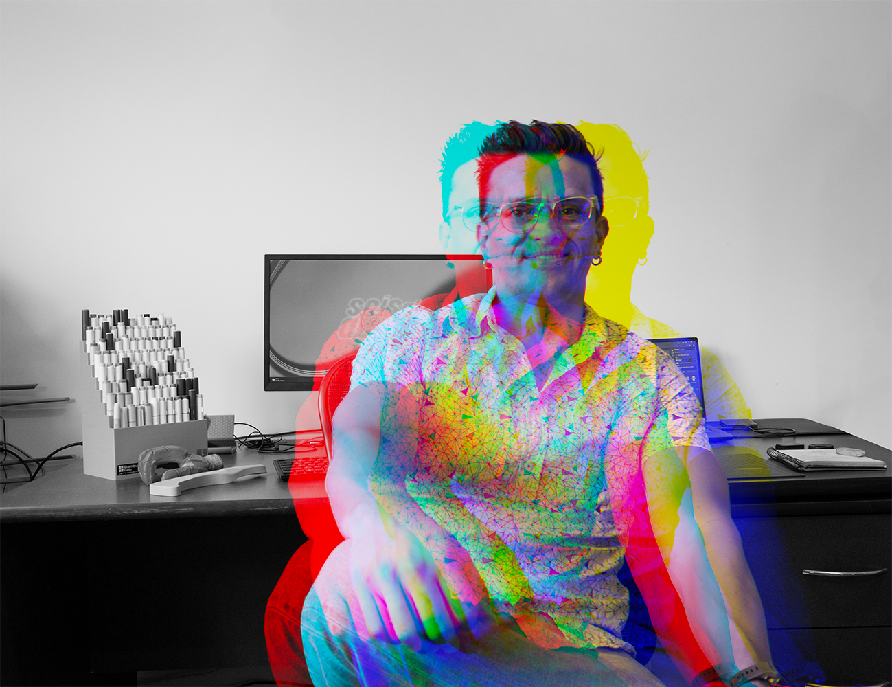 A man with glasses sitting in an office chair at a desk, with a desktop computer and office supplies, overlaid with a colorful glitch effect.