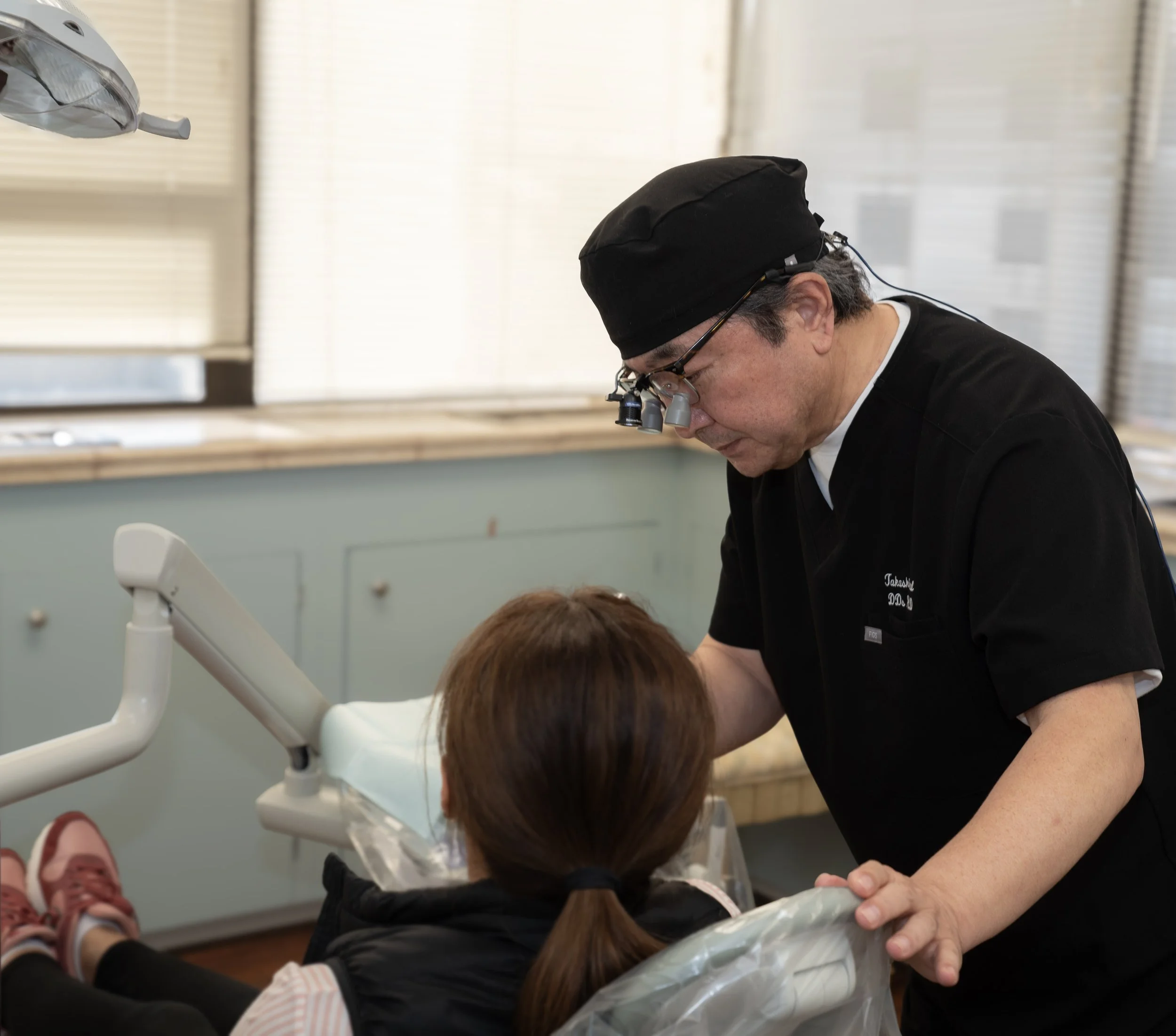 A dentist wearing magnifying loupes examines a patient in a dental clinic with dental equipment and windows with blinds in the background.