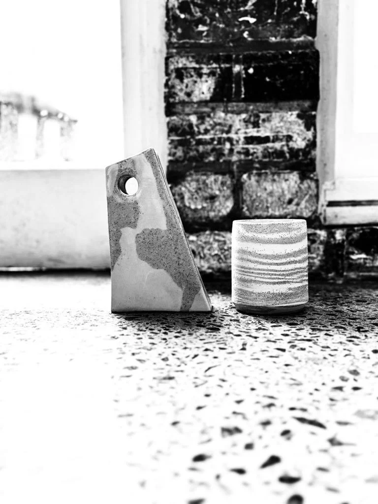 Close-up of a geometric ceramic pitcher and a small striped ceramic cup on a speckled countertop with a rustic brick wall in the background, black and white photo.