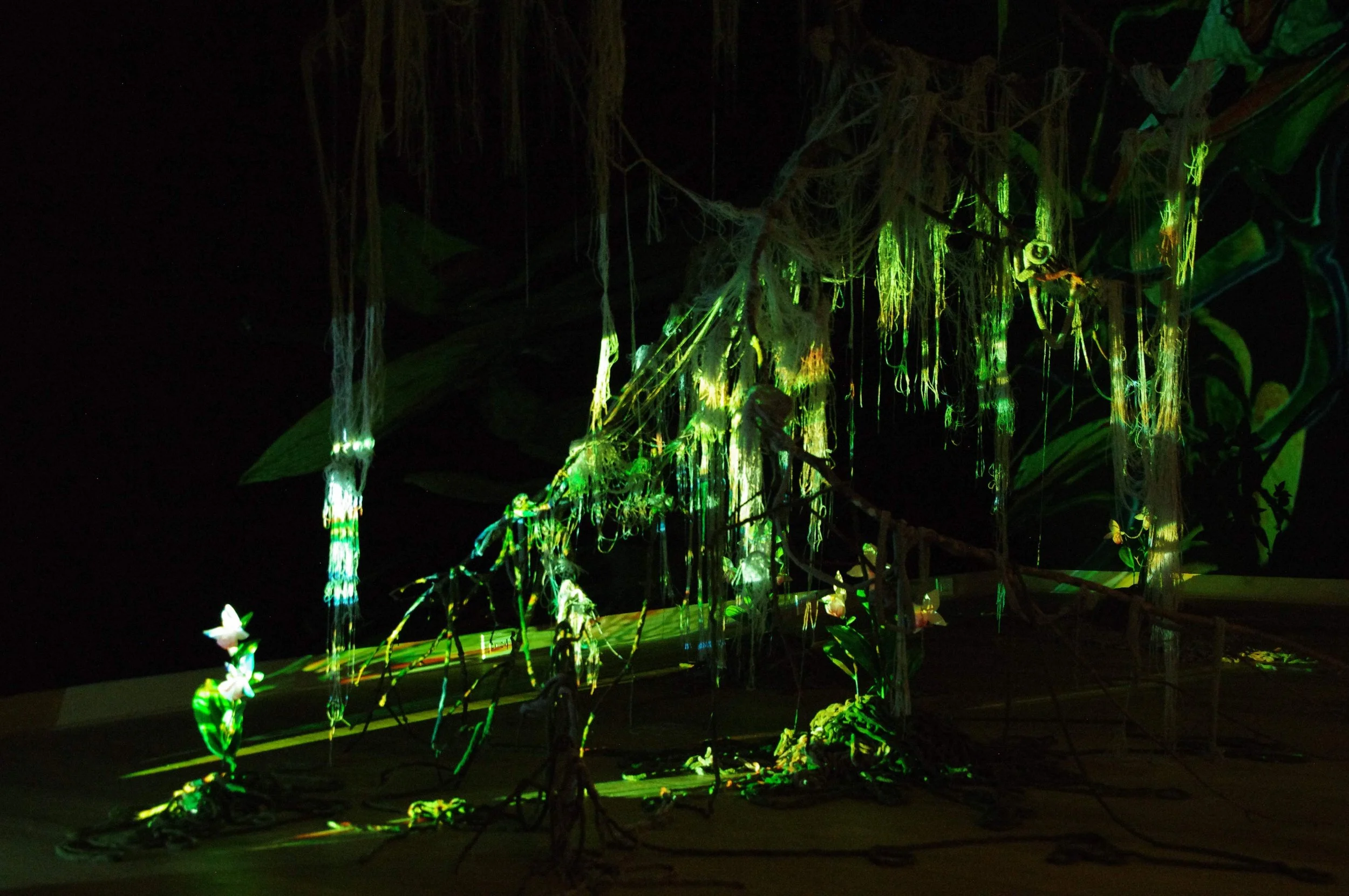 An art installation or display featuring hanging vines and flowers illuminated with green and yellow lights against a dark background.
