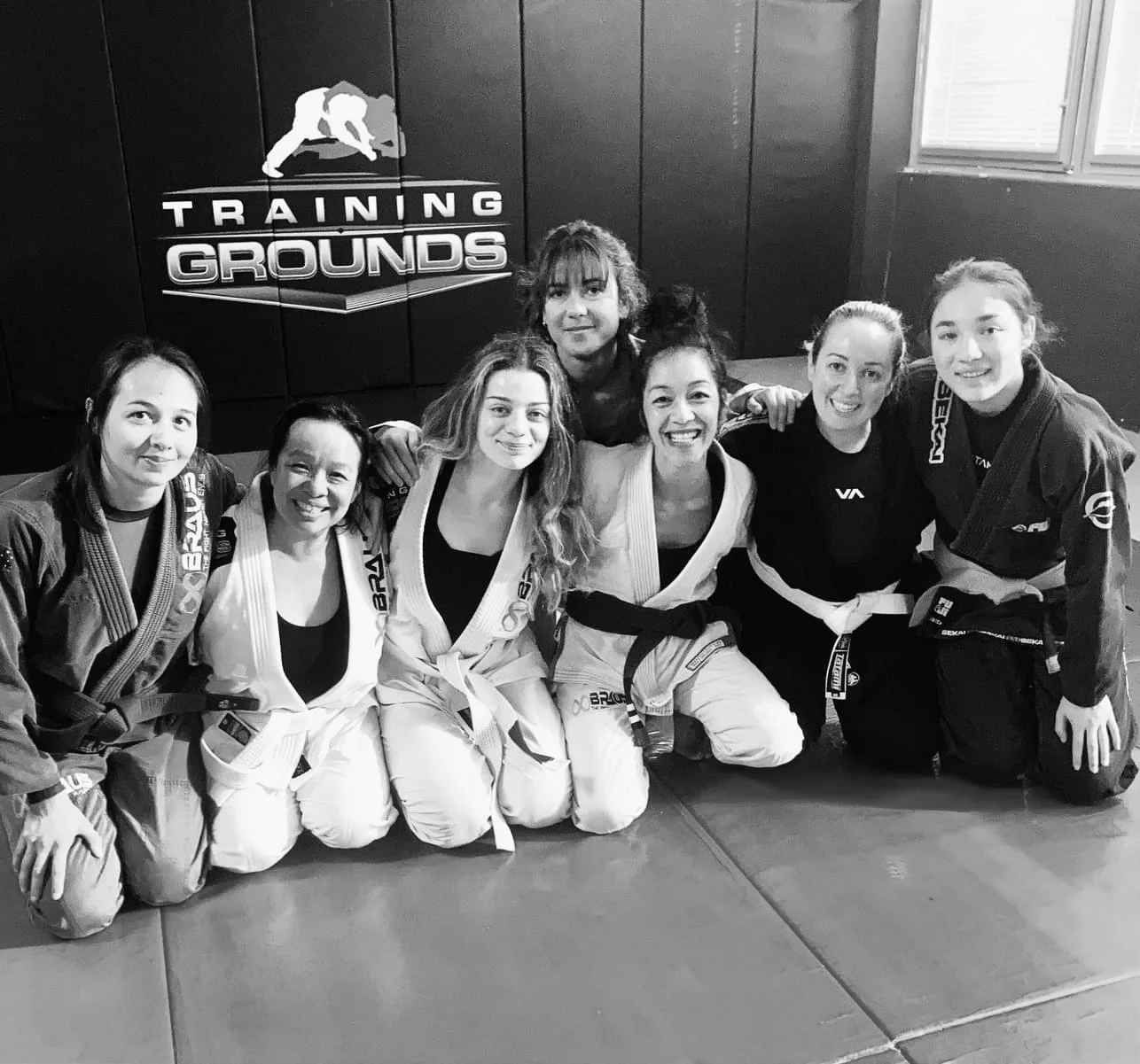 Group of six women practicing Brazilian Jiu-Jitsu in a training gym, kneeling on mats in front of a wall with a logo that reads 'Training Grounds' and features a figure performing a grappling move.