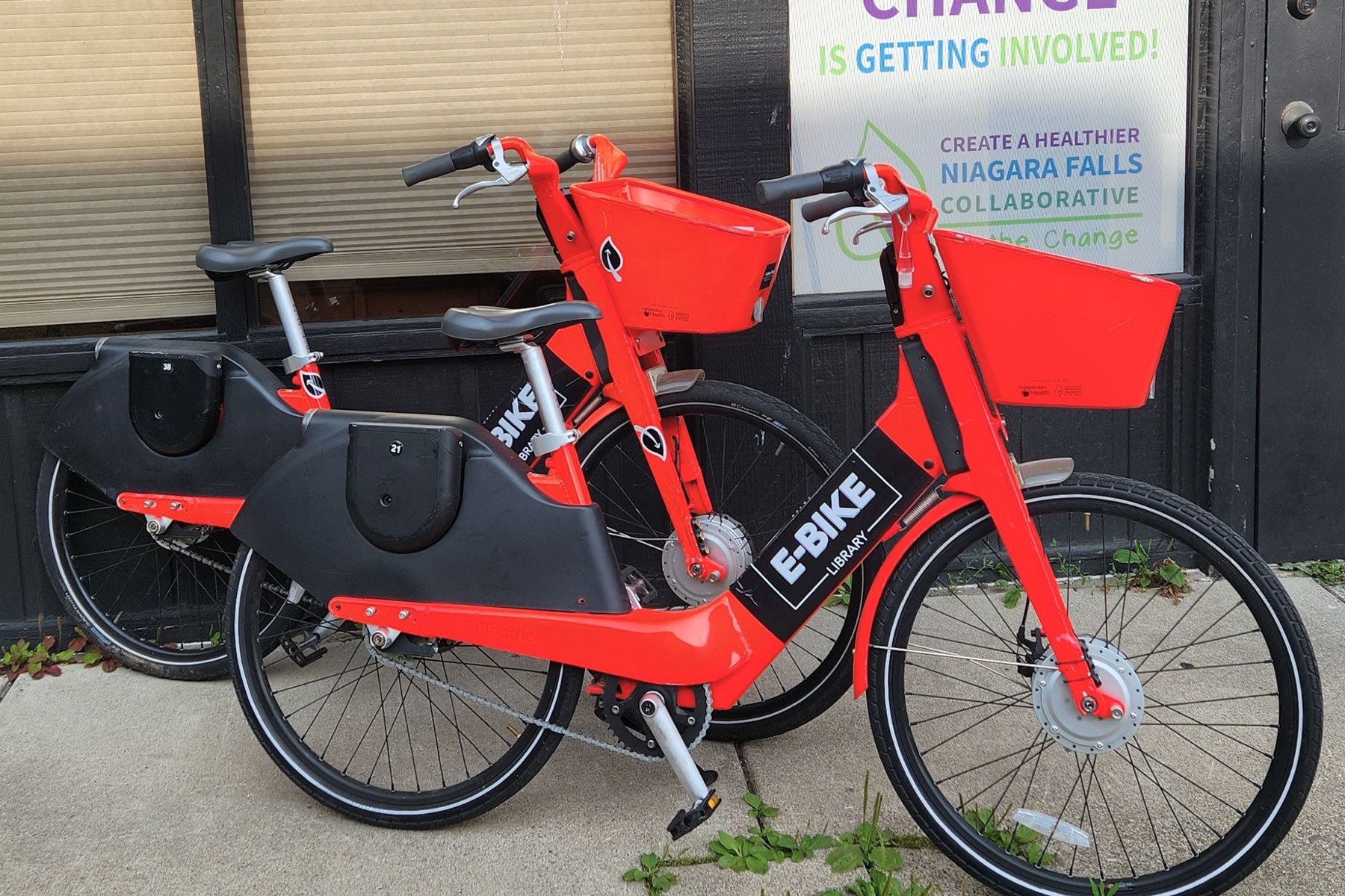 Red and black electric bikeshare bike with front and rear baskets, parked outside a building.