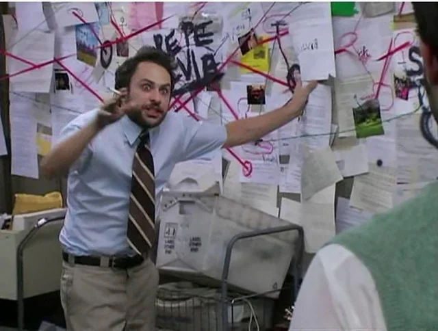 Man in office pointing to wall covered with papers, photos, and red string connecting various items in a conspiracy or investigation scene.