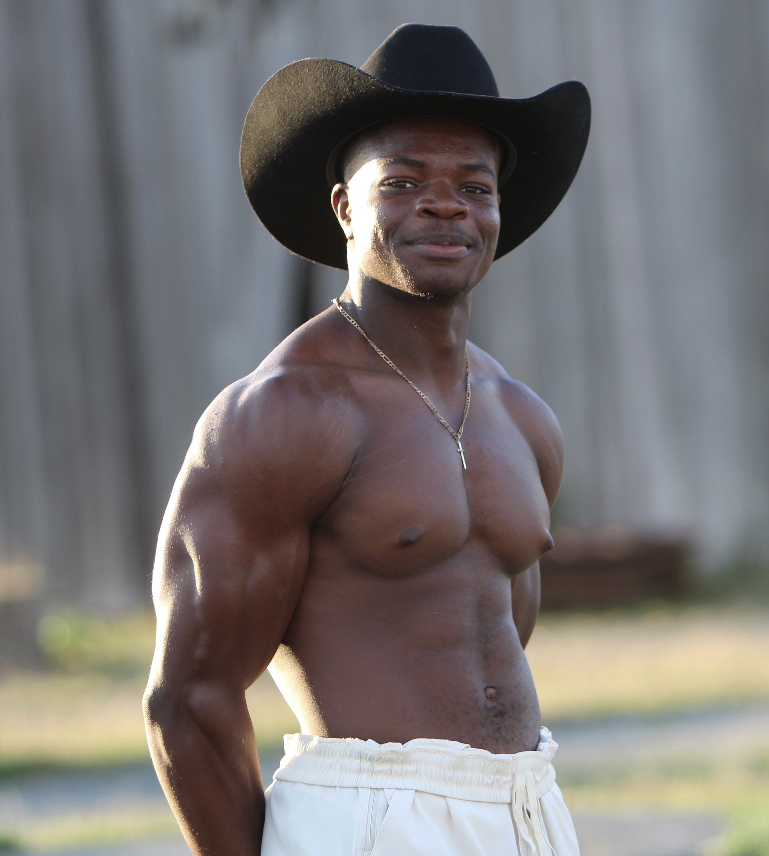 A shirtless man wearing a large black cowboy hat, white pants, and a cross necklace, standing outdoors in front of a wooden fence.