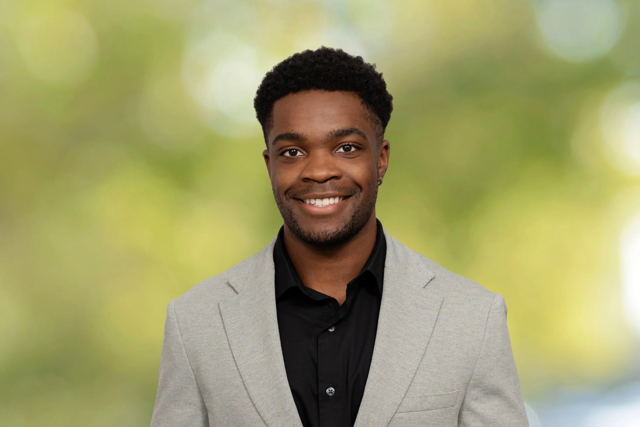A young man with dark hair and a beard smiling, wearing a light gray blazer and black shirt, standing outdoors with a blurred green background.