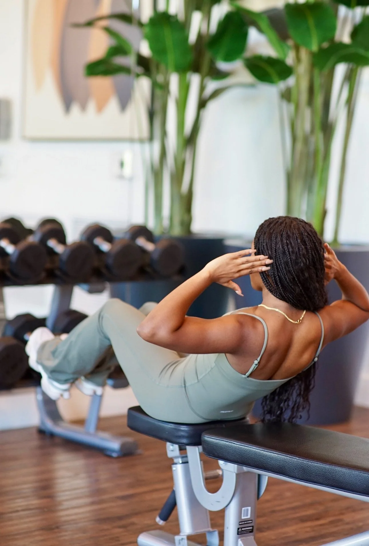 A woman with braided hair doing crunches on a workout bench in a gym, with a row of dumbbells and large green plants in the background.