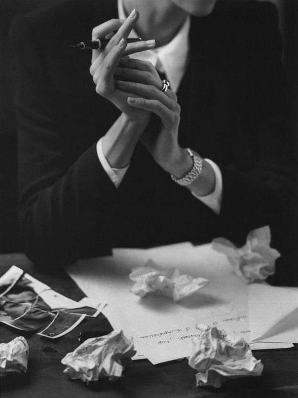 A person in a suit holding a cigarette with crumpled papers and a sheet of paper on a desk.